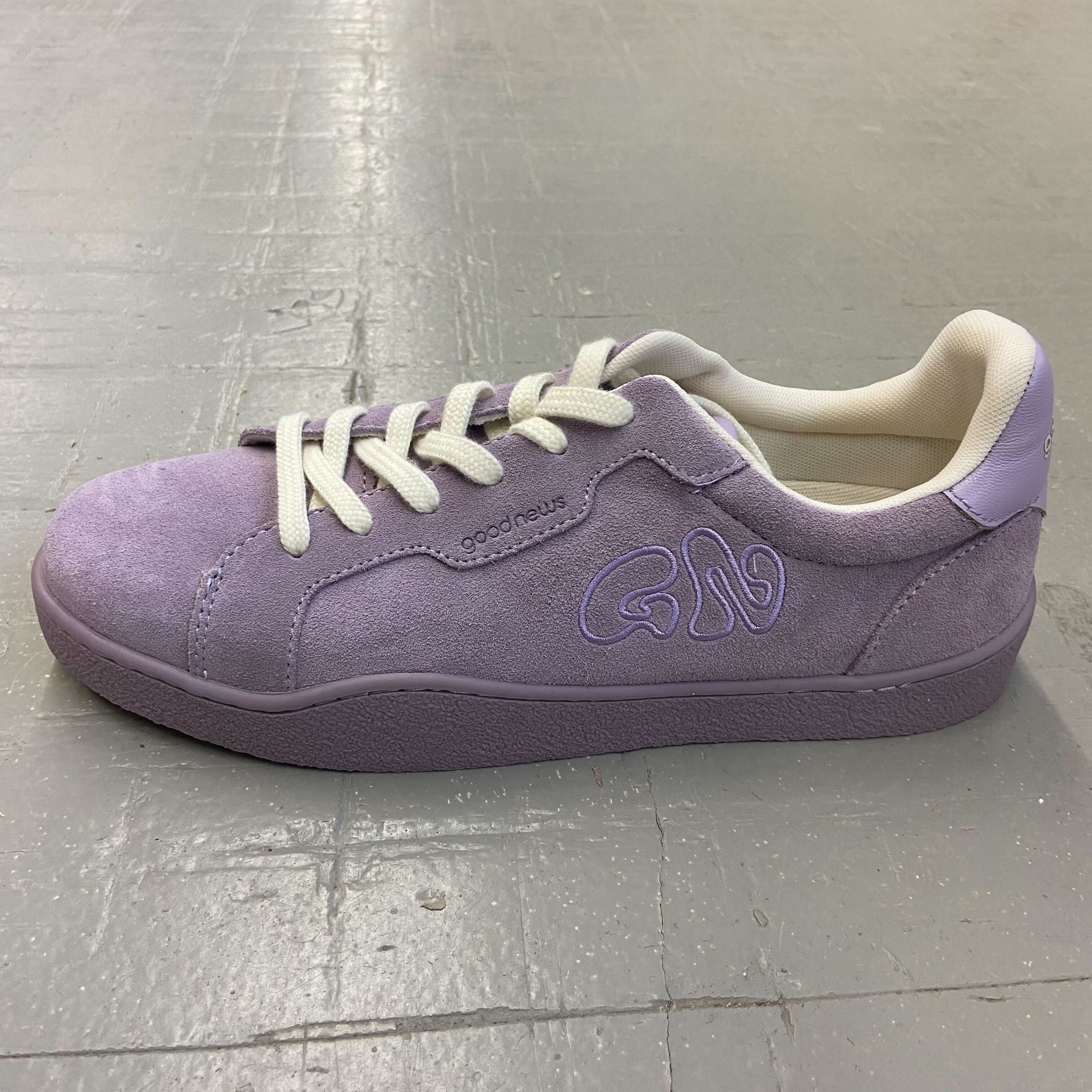 Lila Wildledersneaker mit weißen Schnürsenkeln und gesticktem Logo, auf grauem Boden platziert.