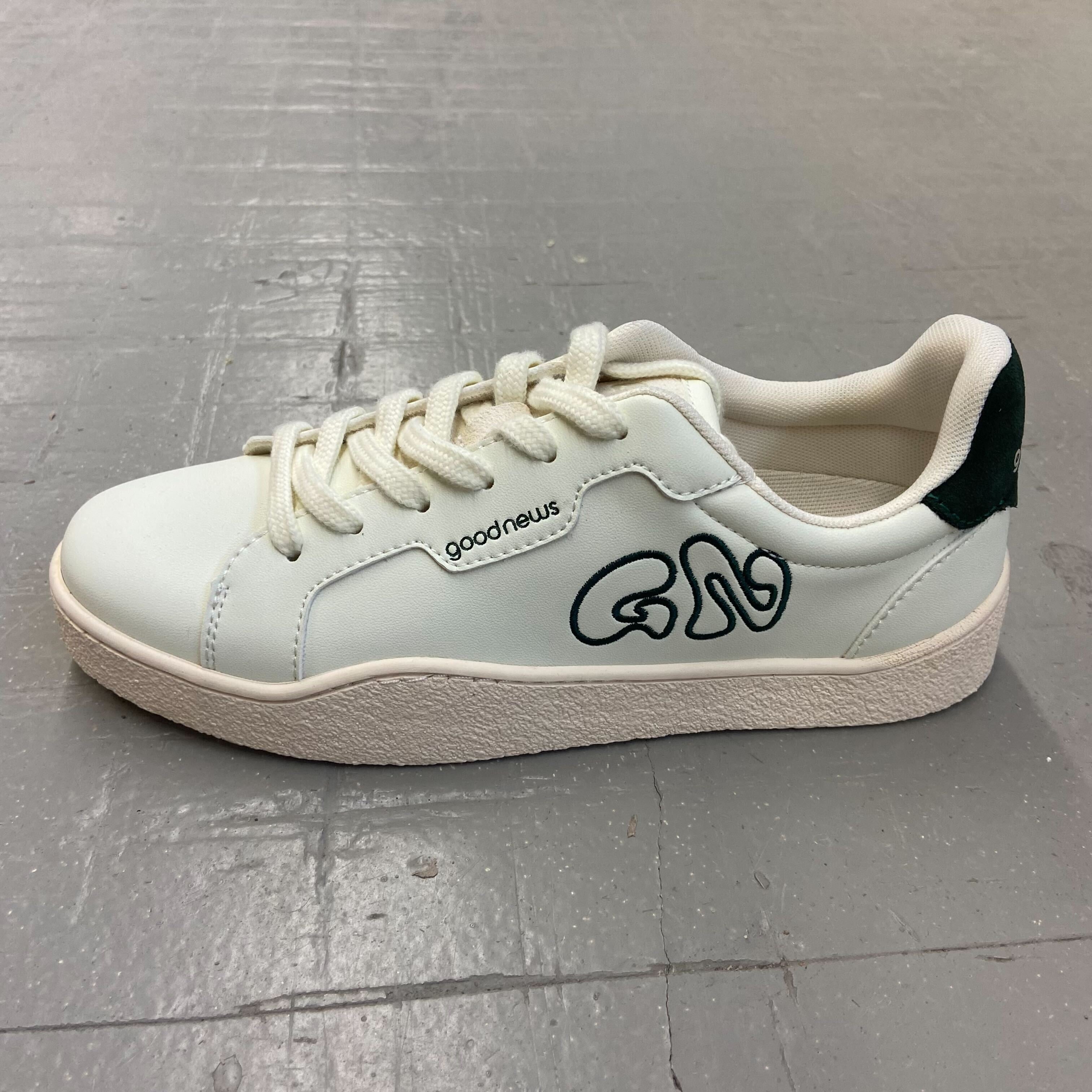 Weißer Sneaker mit dunkelgrünem Logo, der auf einem grauen Boden steht.