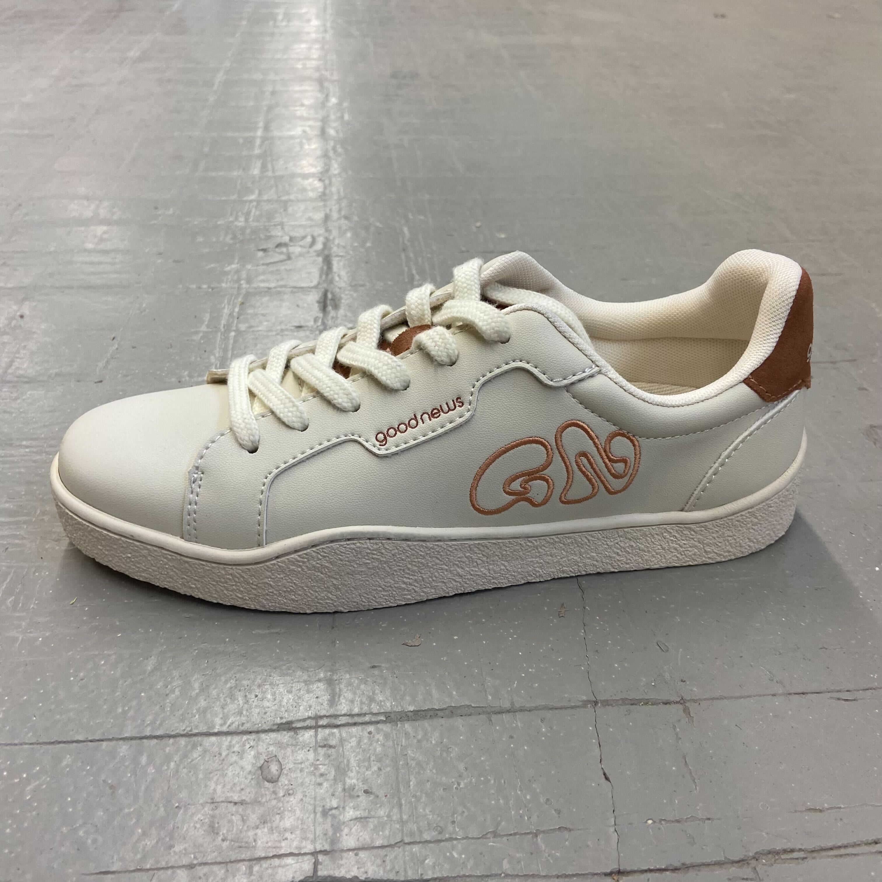 Weiße Sneaker mit dem Schriftzug "goodnews" und einem braunen Akzent an der Ferse, auf grauem Boden.