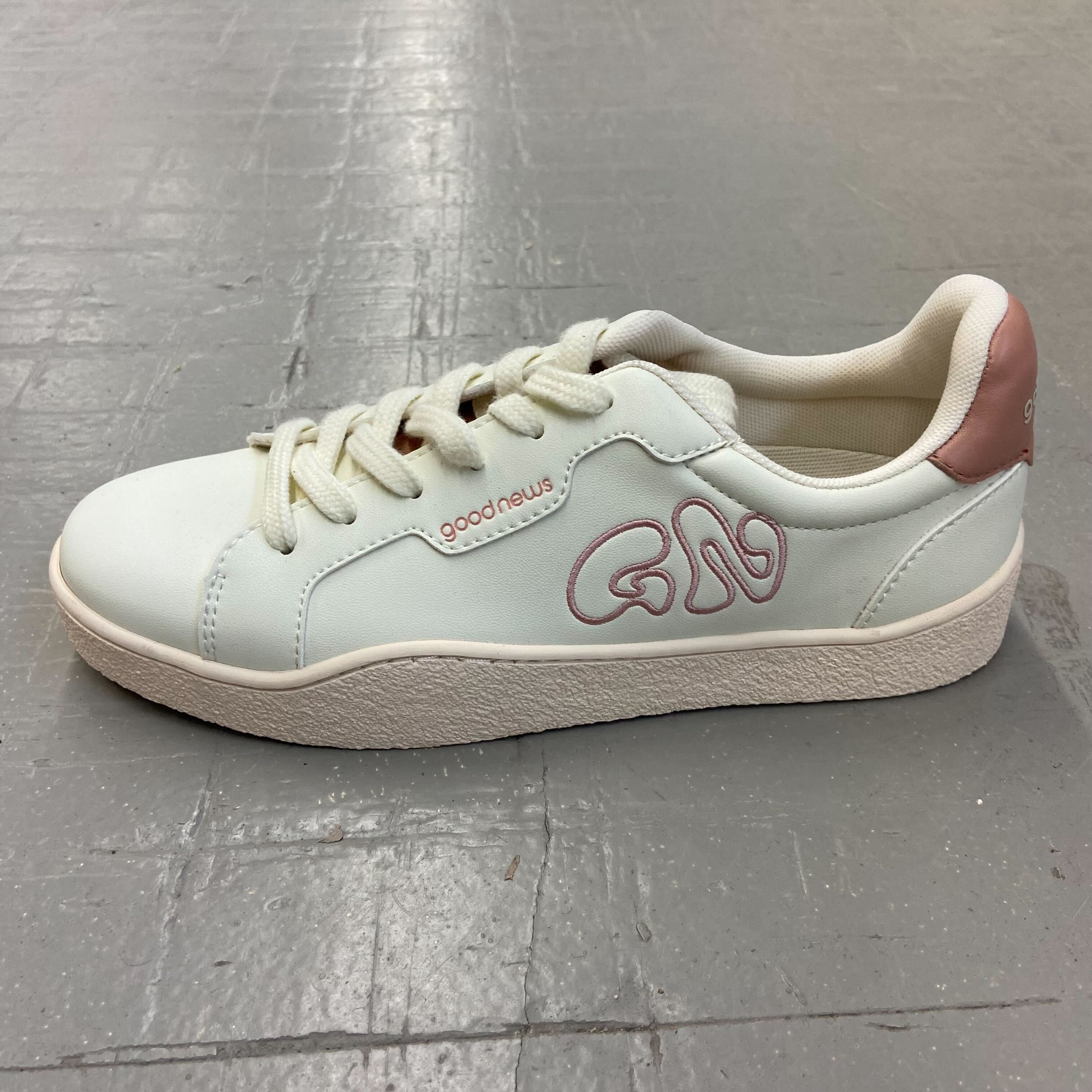Weißer Sneaker mit rosa Details und der Aufschrift "good news" auf grauem Boden platziert.
