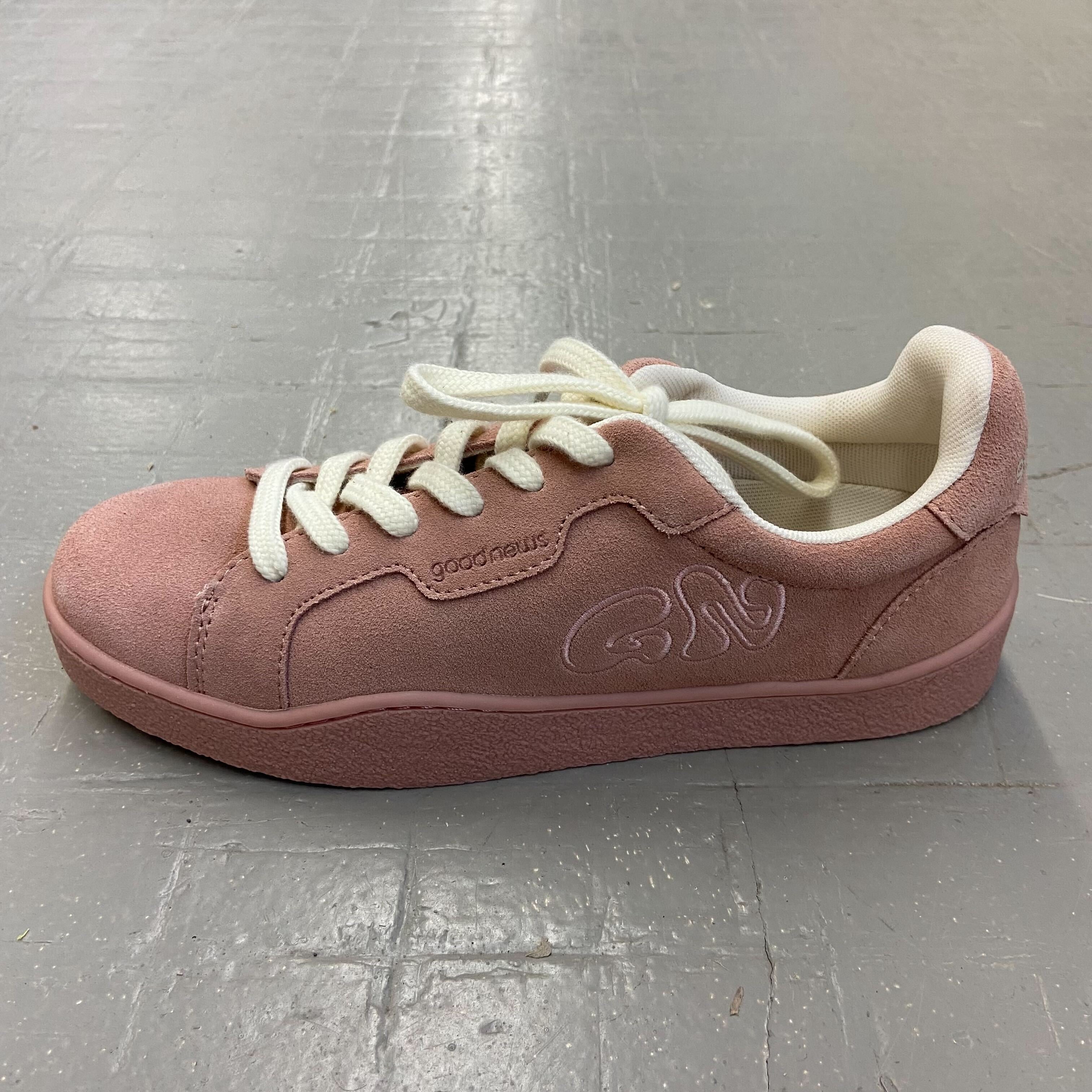 Rosa Sneaker mit weißen Schnürsenkeln auf grauem Boden, mit aufgesticktem "Good News"-Logo an der Seite.