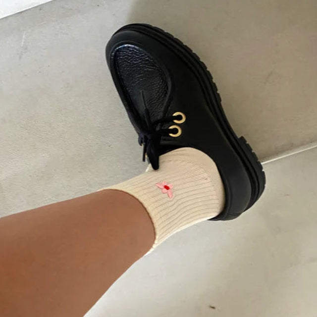 Eine Person, die cremefarbene Socken mit einem kleinen roten Herz und schwarze Lederschuhe mit goldenen Ösen trägt, steht auf einem hellen Boden.