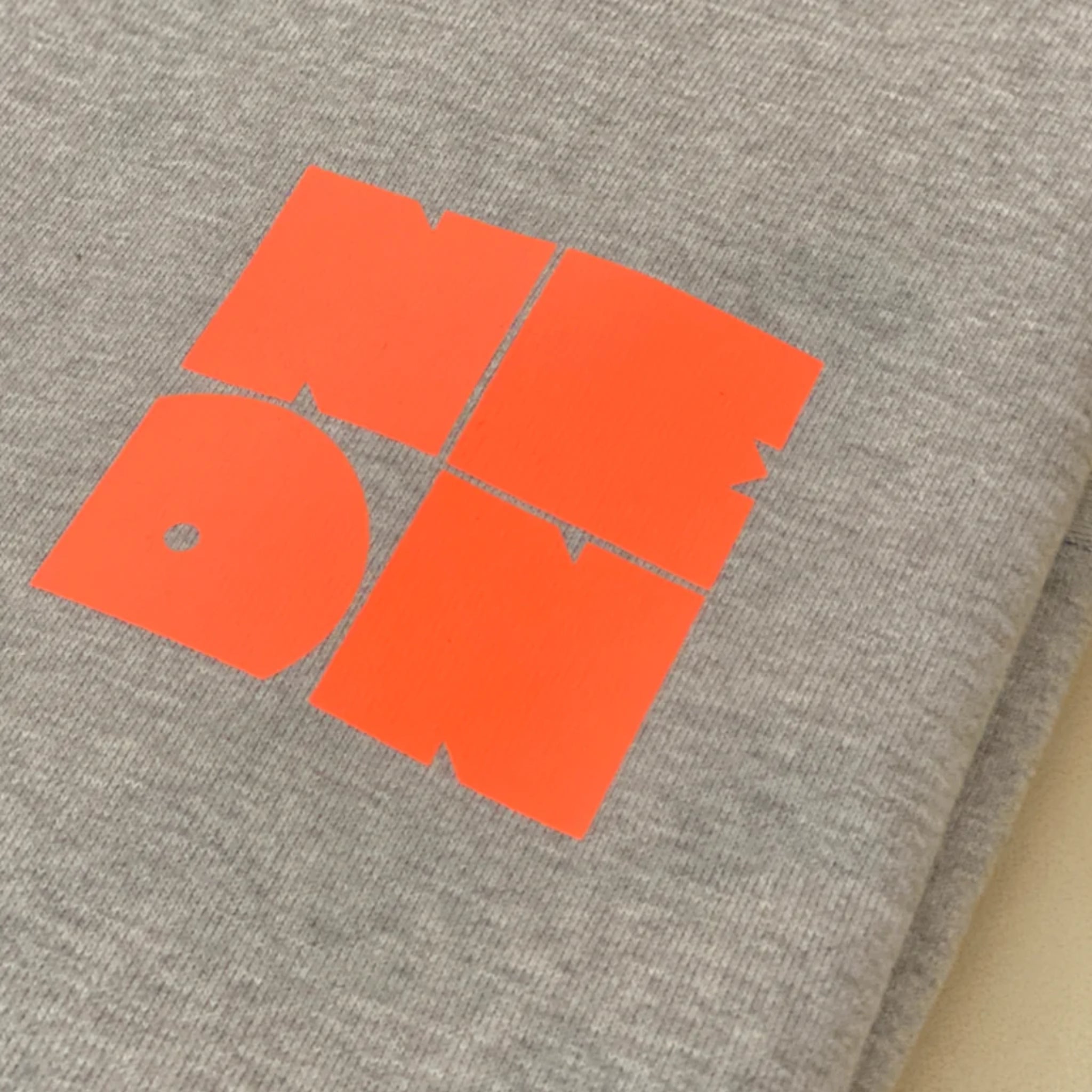 Nahaufnahme eines grauen Stoffes, möglicherweise eines Sweatshirts, mit einer auffälligen, blockartigen orangefarbenen Grafik, die in vier Quadrate mit den Buchstaben ND, DN aufgeteilt ist, die in einem Gittermuster angeordnet sind.