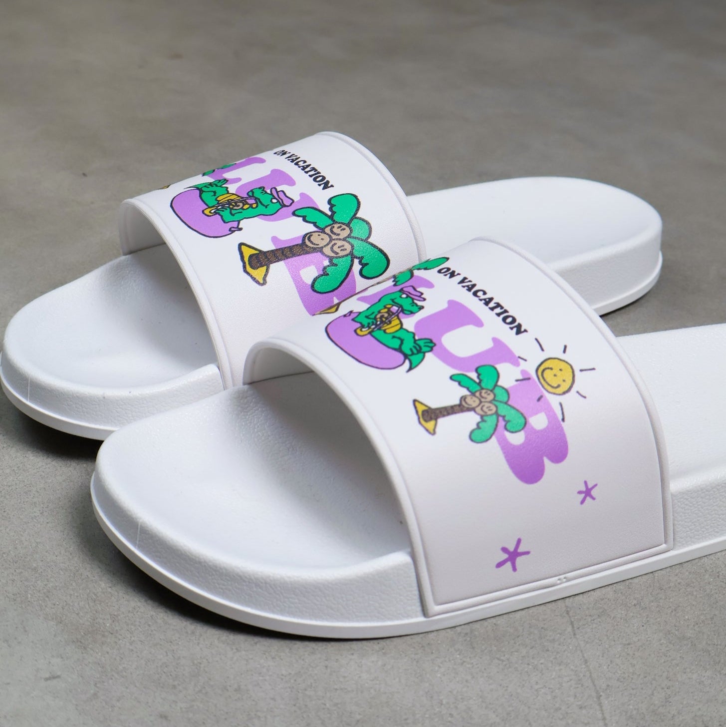 Ein Paar weiße Slide-Sandalen auf grauem Untergrund, mit bunten Cartoon-Motiven wie Palmen, Trompeten, einer Sonne und der Aufschrift ON VACATION auf den Riemen.