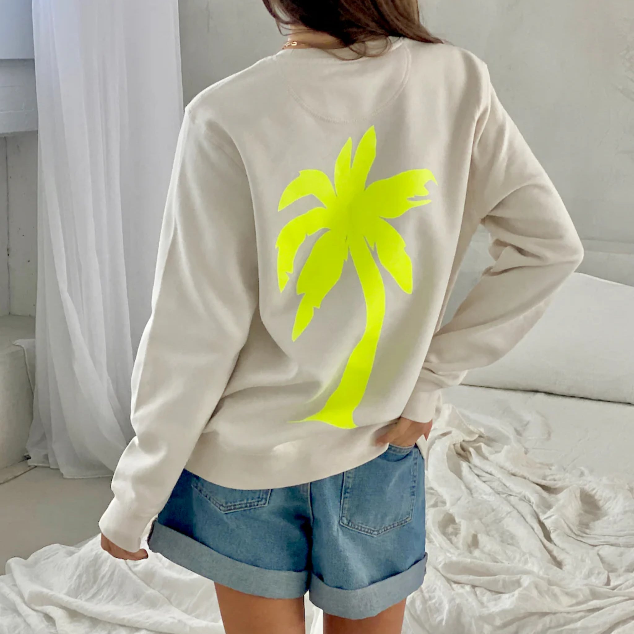 Eine Person, die ein hellbeiges Sweatshirt mit einer großen gelben Palmengrafik auf dem Rücken und Jeansshorts trägt, steht in einem sanft beleuchteten, minimalistischen Schlafzimmer mit weißer Bettwäsche.