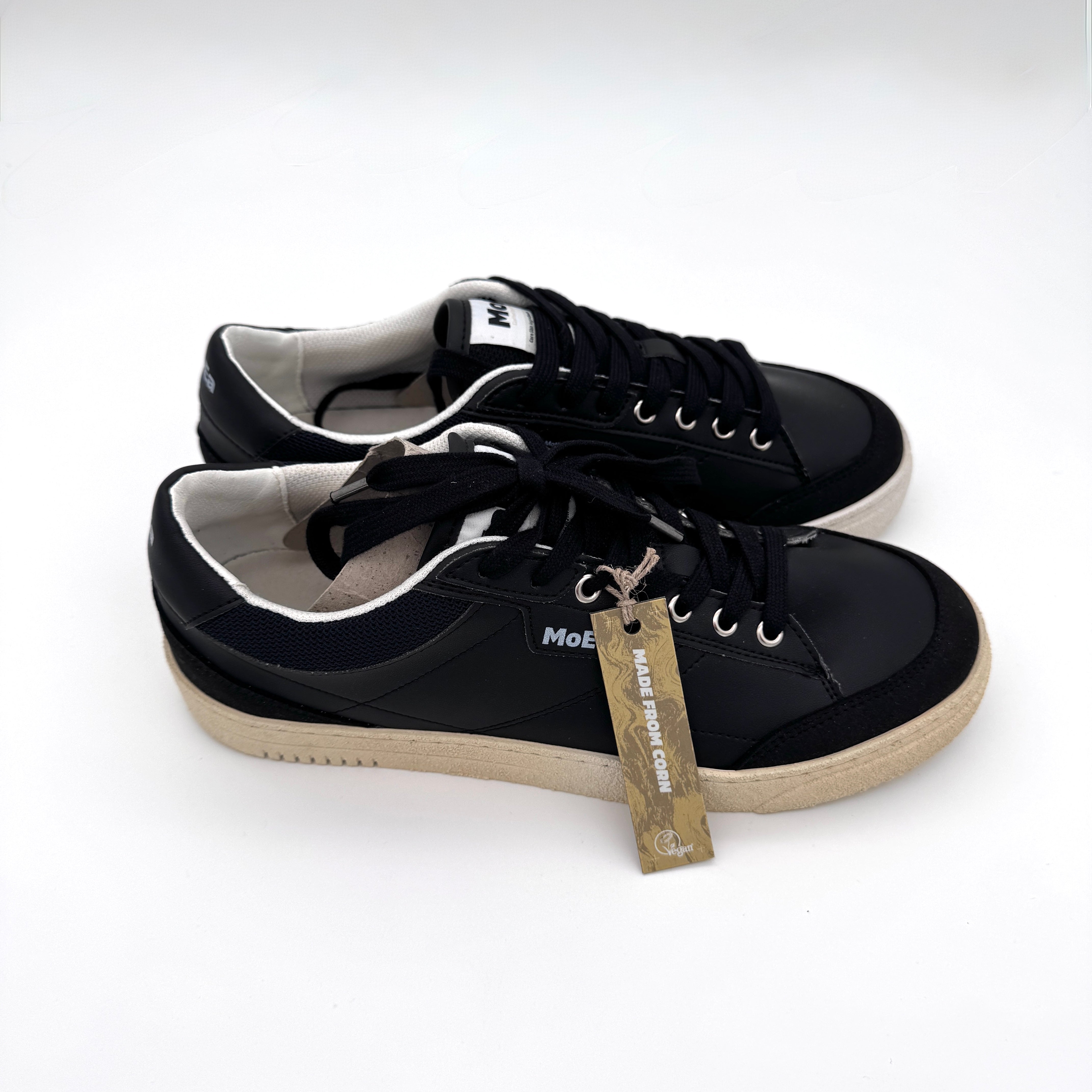 Ein Paar schwarzer Turnschuhe mit weißer Sohle und schwarzen Schnürsenkeln, mit einem beigefarbenen Schildchen am rechten Schuh, das auf einem schlichten weißen Hintergrund angebracht ist.