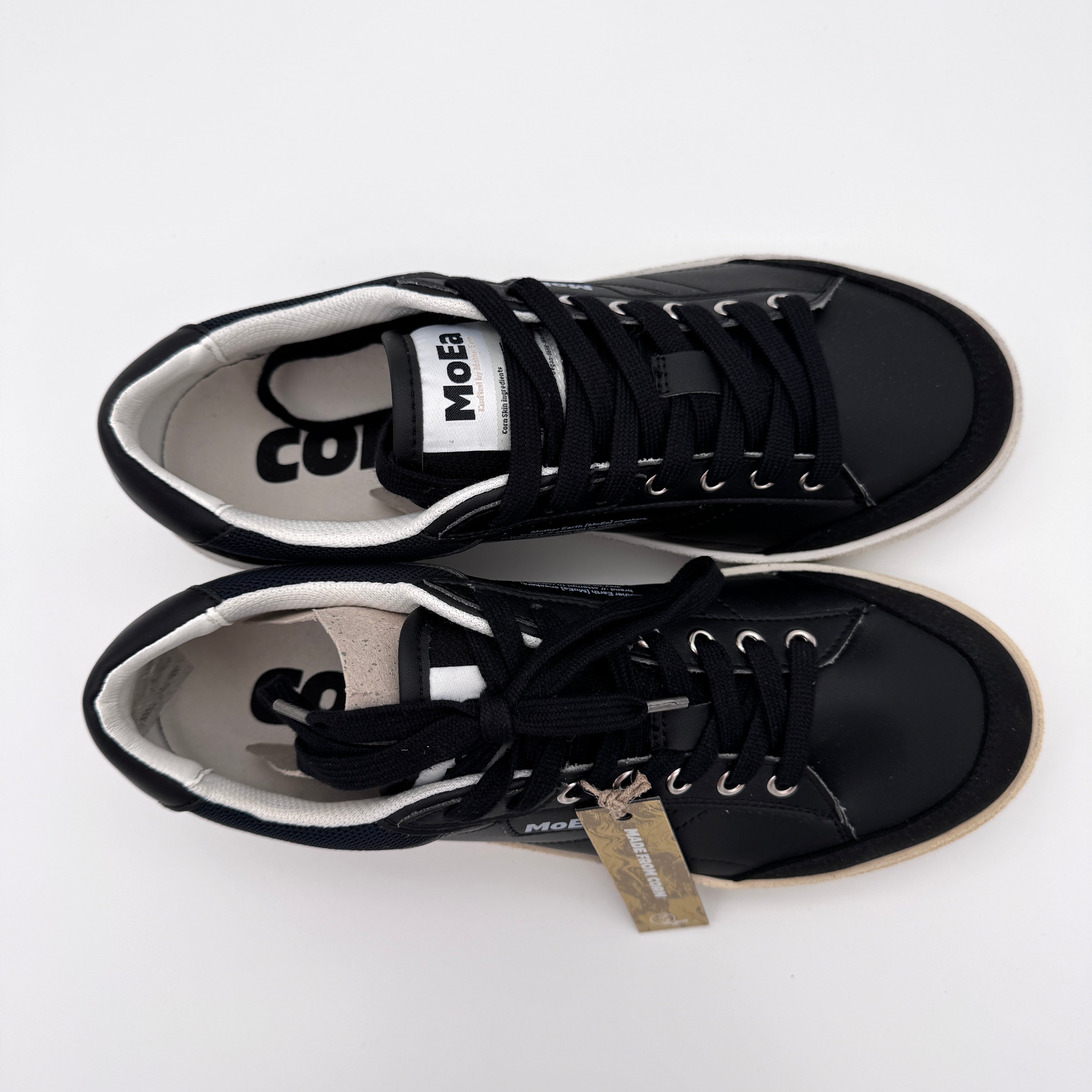 Ein Paar schwarze Turnschuhe mit weißer Sohle, weißem Innenfutter und schwarzen Schnürsenkeln stehen nebeneinander auf einem weißen Hintergrund. An den Schnürsenkeln des rechten Schuhs ist ein braunes Etikett befestigt.