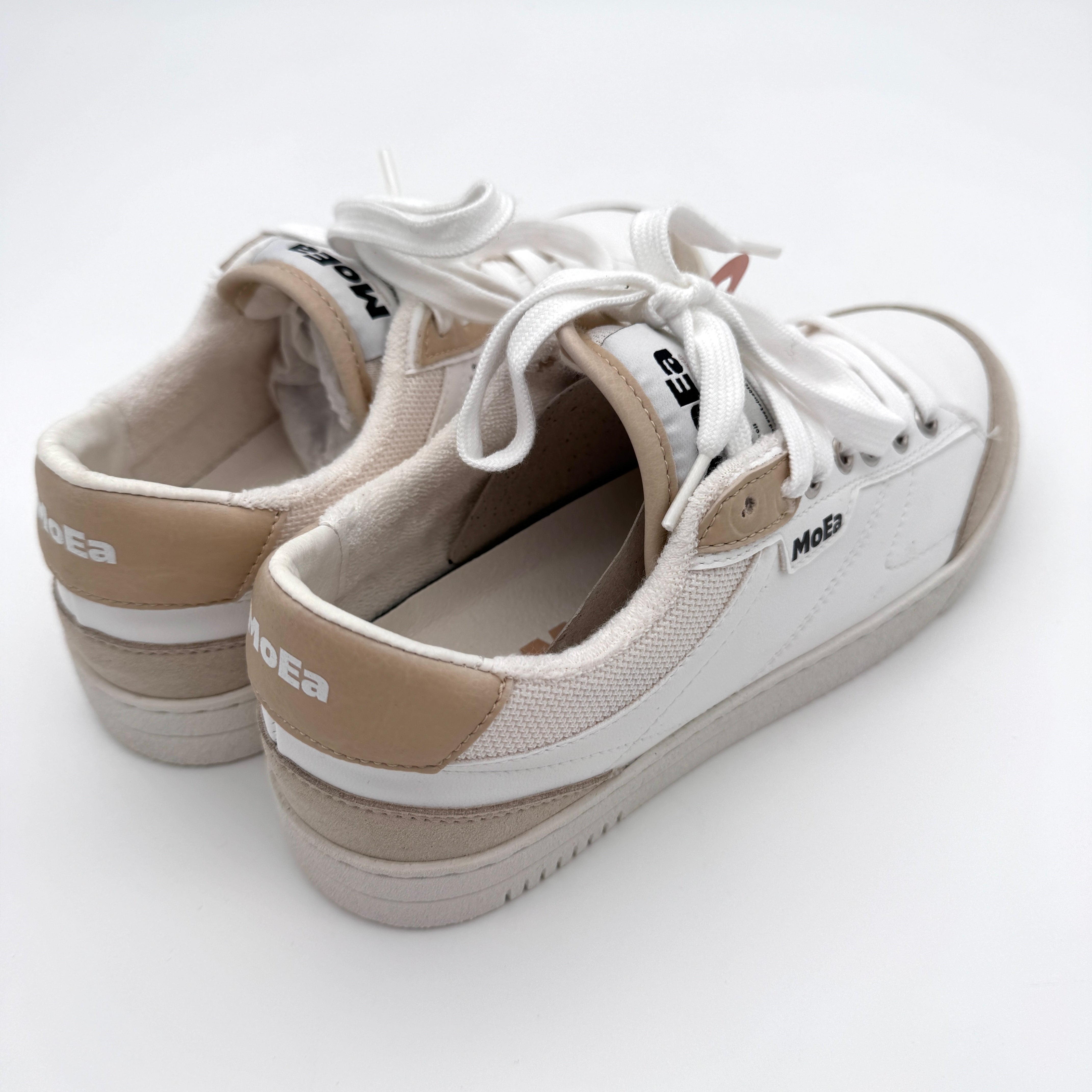 Ein Paar weiße und beigefarbene Turnschuhe mit weißen Schnürsenkeln, mit dem Markennamen MOEA an der Ferse und an der Seite, auf einem weißen Hintergrund.