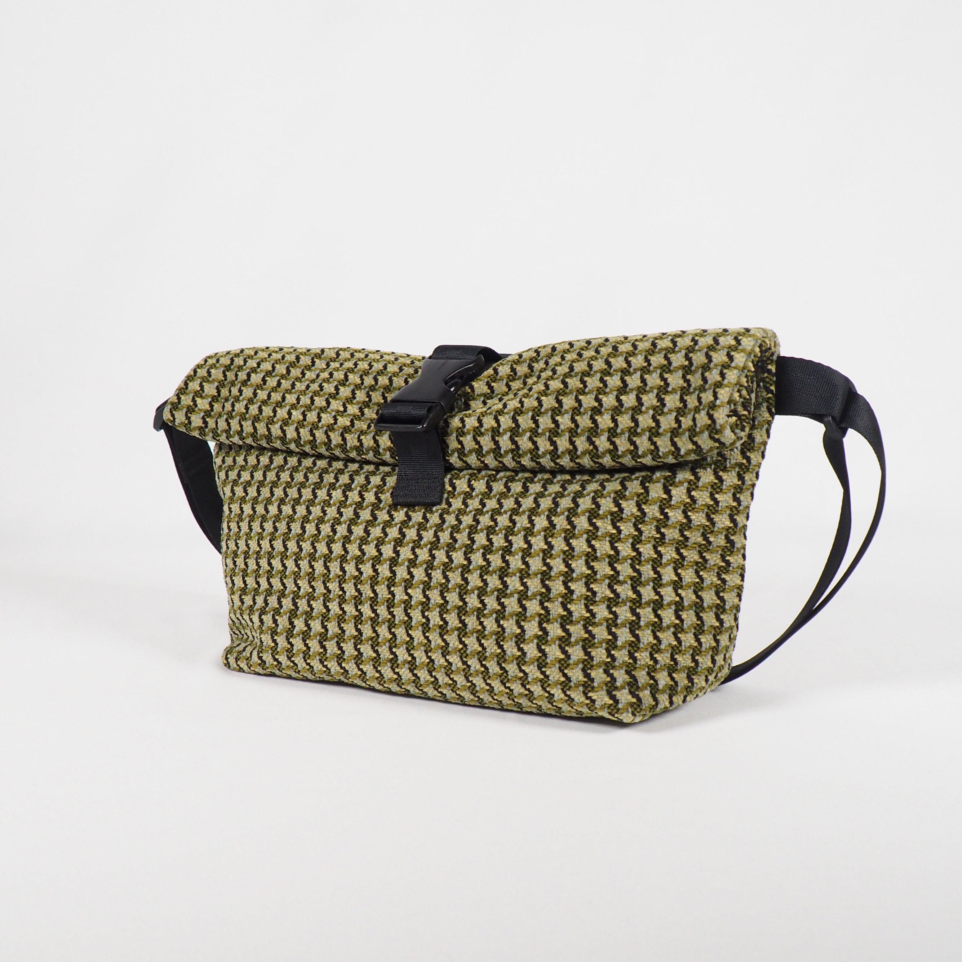 Eine strukturierte Crossbody-Tasche mit grünem und schwarzem Webmuster, einem schwarzen verstellbaren Riemen und einem schwarzen Schnallenverschluss auf der Vorderseite vor einem schlichten weißen Hintergrund.