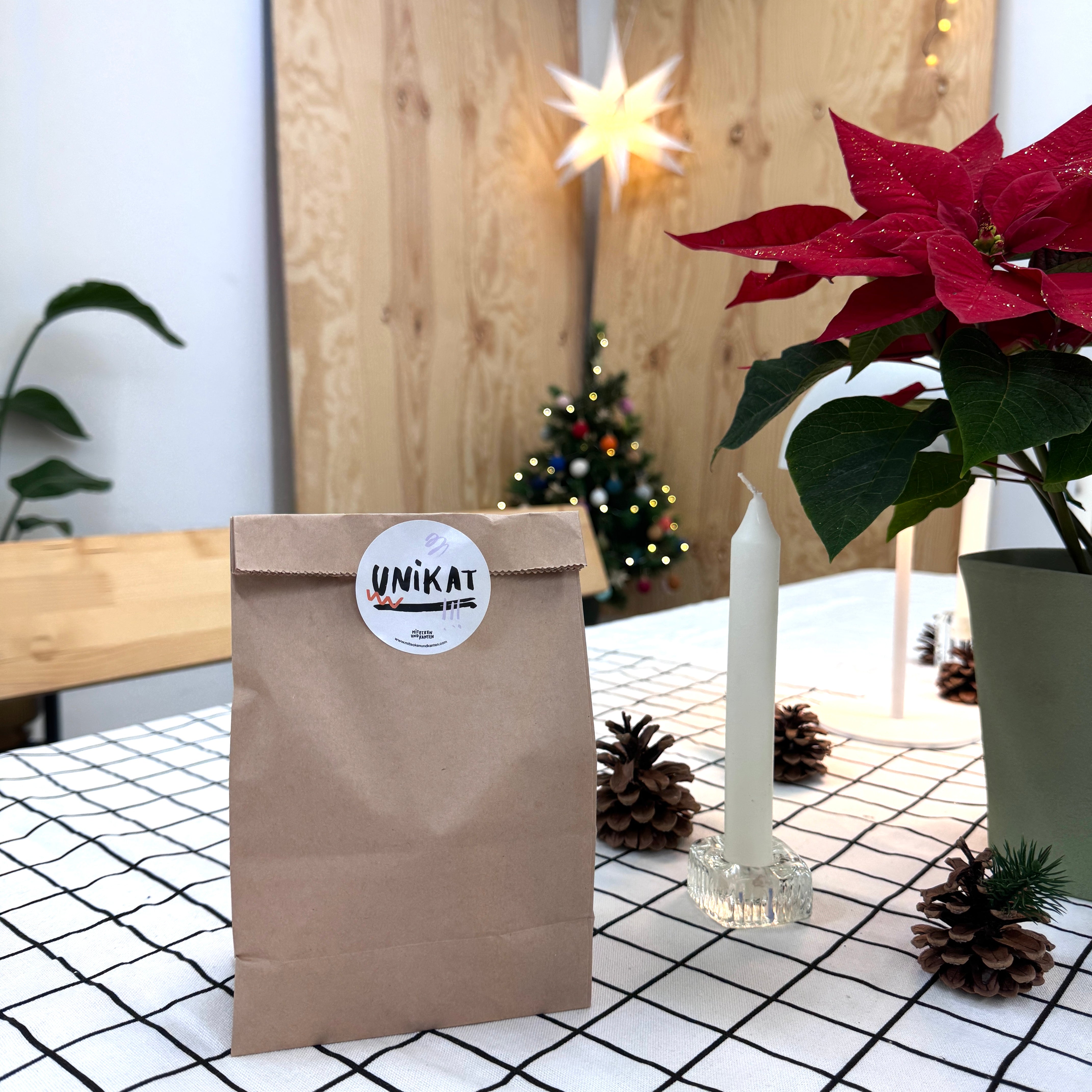Eine braune Papiertüte mit einem UNIKAT-Aufkleber liegt auf einem karierten Tischtuch neben einer weißen Kerze, Tannenzapfen und einem getopften Weihnachtsstern. Im Hintergrund stehen ein kleiner Weihnachtsbaum und eine Sternendekoration.