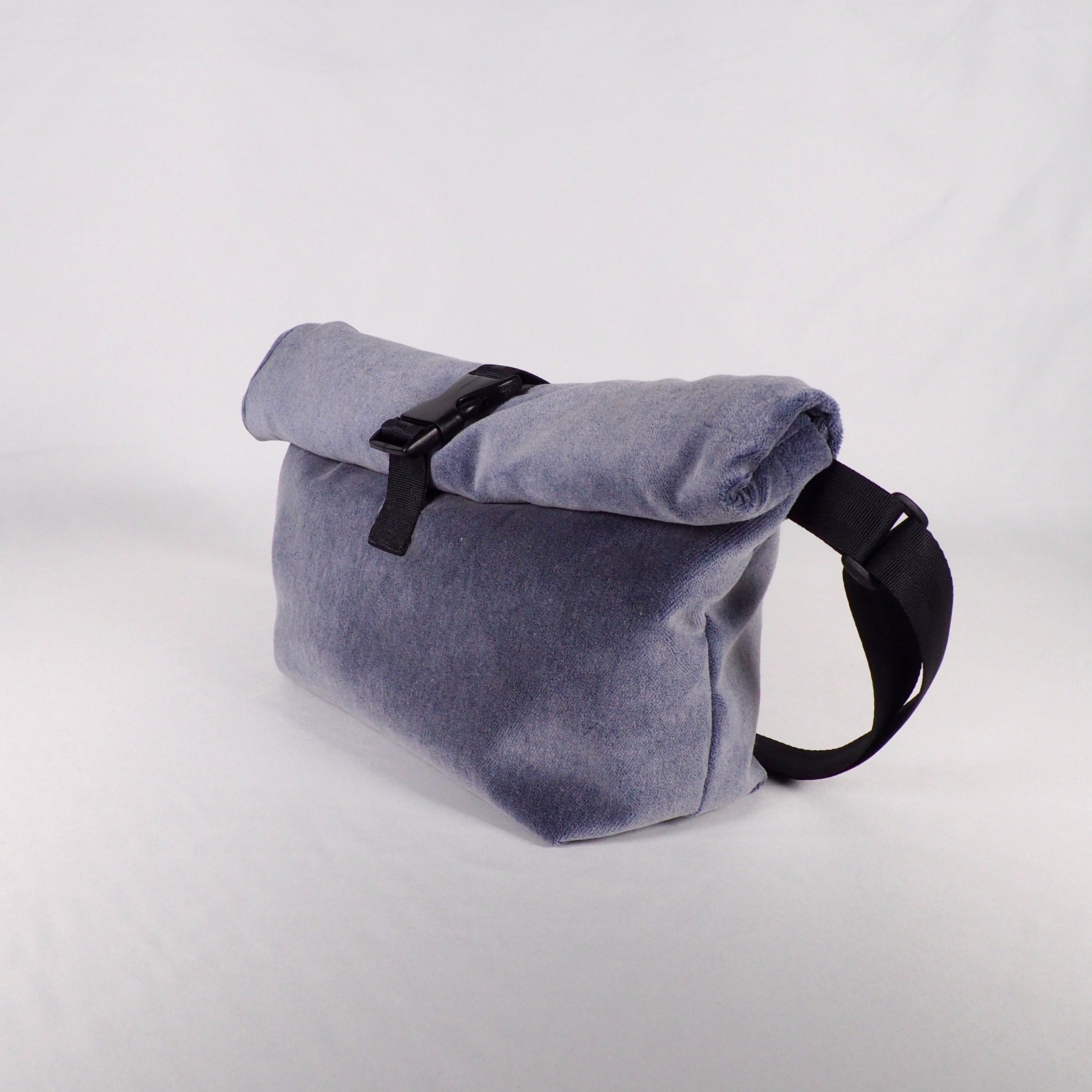 Eine hellgraue Rolltop-Tasche aus Stoff mit einem schwarzen Schnallenverschluss und einem verstellbaren schwarzen Riemen ist auf einem schlichten weißen Hintergrund abgebildet.