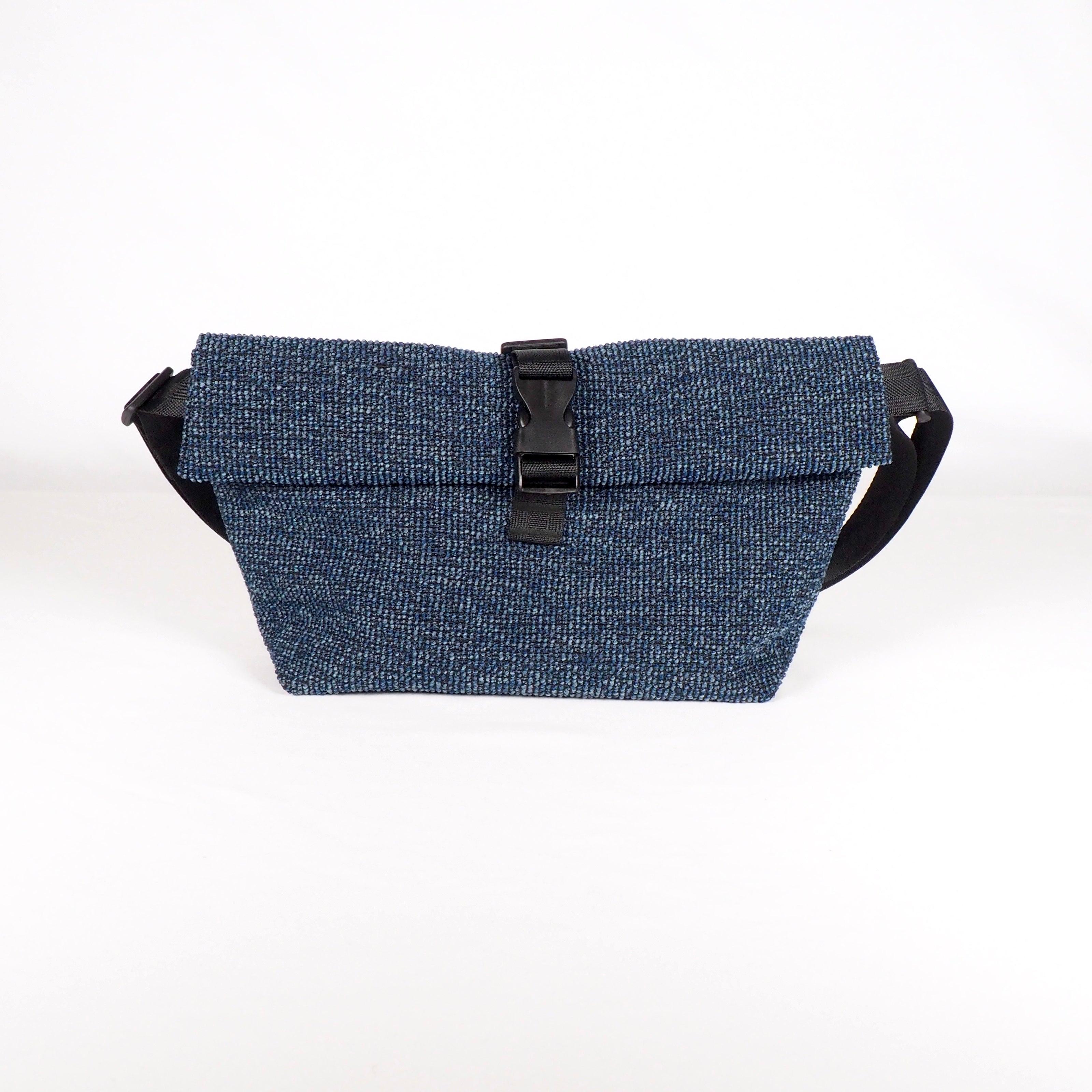 Eine blaue Crossbody-Tasche aus strukturiertem Stoff mit einer umklappbaren Klappe, die mit einer schwarzen Kunststoffschnalle gesichert ist, auf weißem Hintergrund.