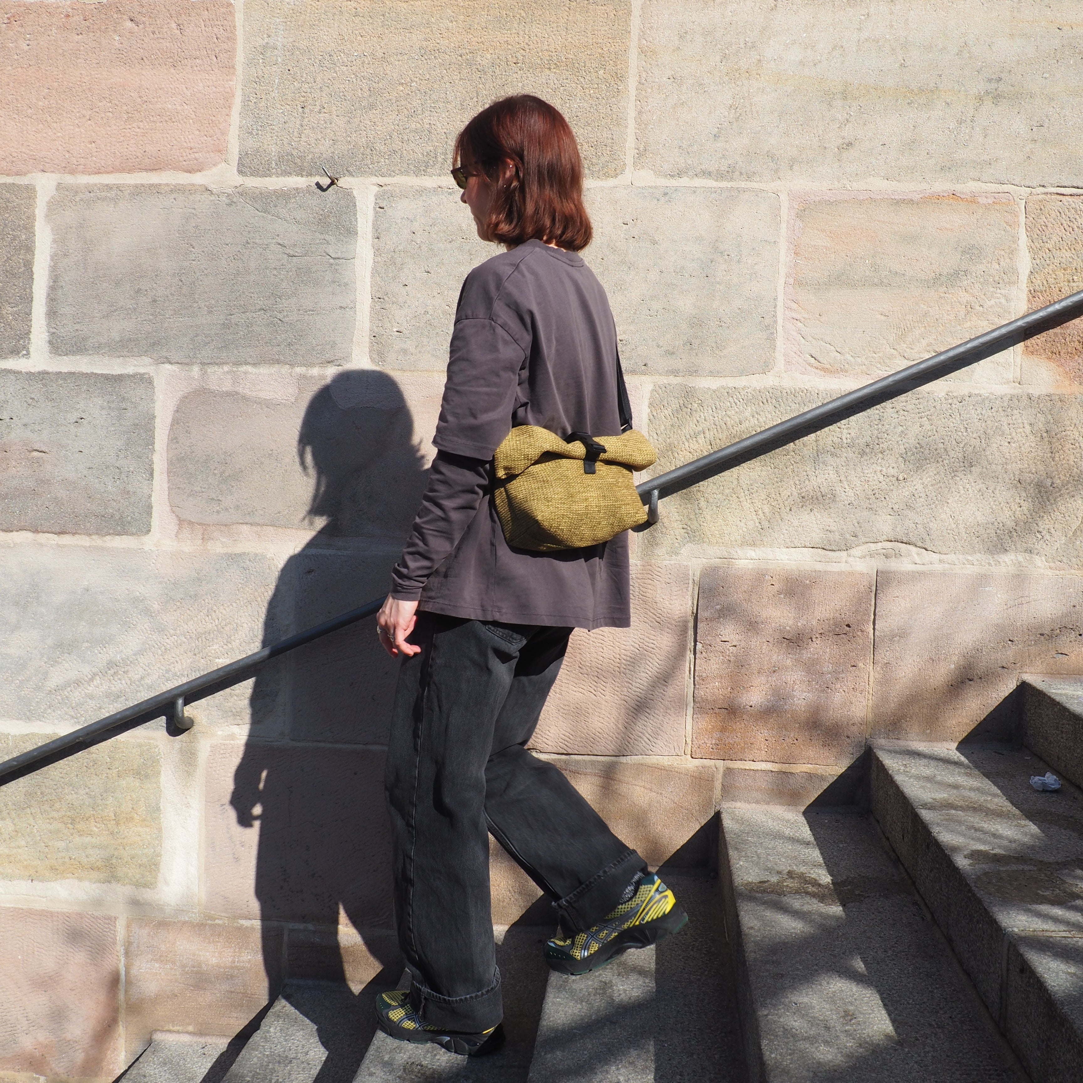 Eine Person mit schulterlangen braunen Haaren, Sonnenbrille und einer gelben Tasche geht an einem sonnigen Tag steinerne Stufen an einer strukturierten Ziegelmauer hinauf.