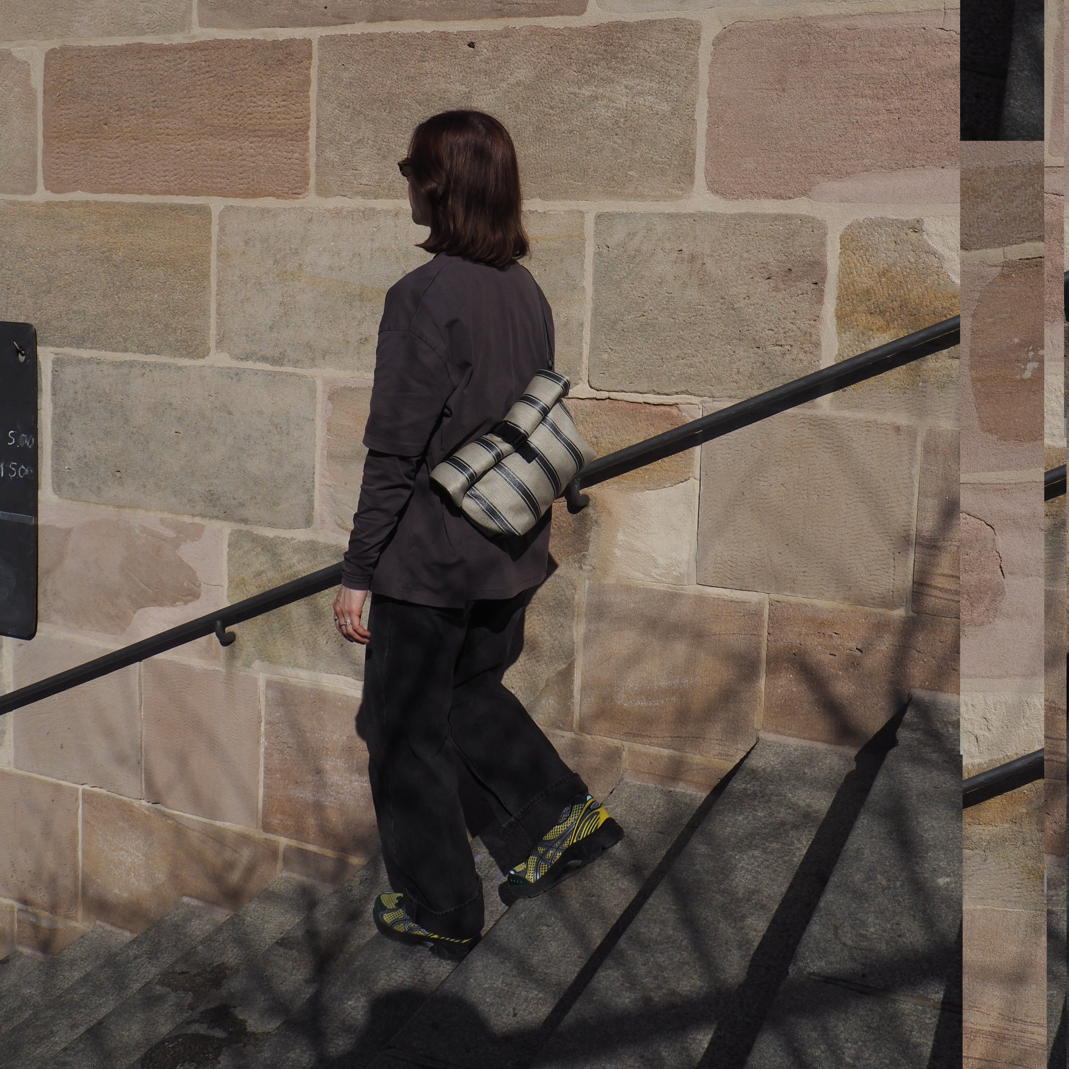 Eine Person mit schulterlangen braunen Haaren, dunkler Kleidung und Turnschuhen geht eine Steintreppe neben einer großen Backsteinmauer hinauf und trägt eine gestreifte Tasche auf dem Rücken. Die Schatten eines Geländers und eines Baumes sind sichtbar.