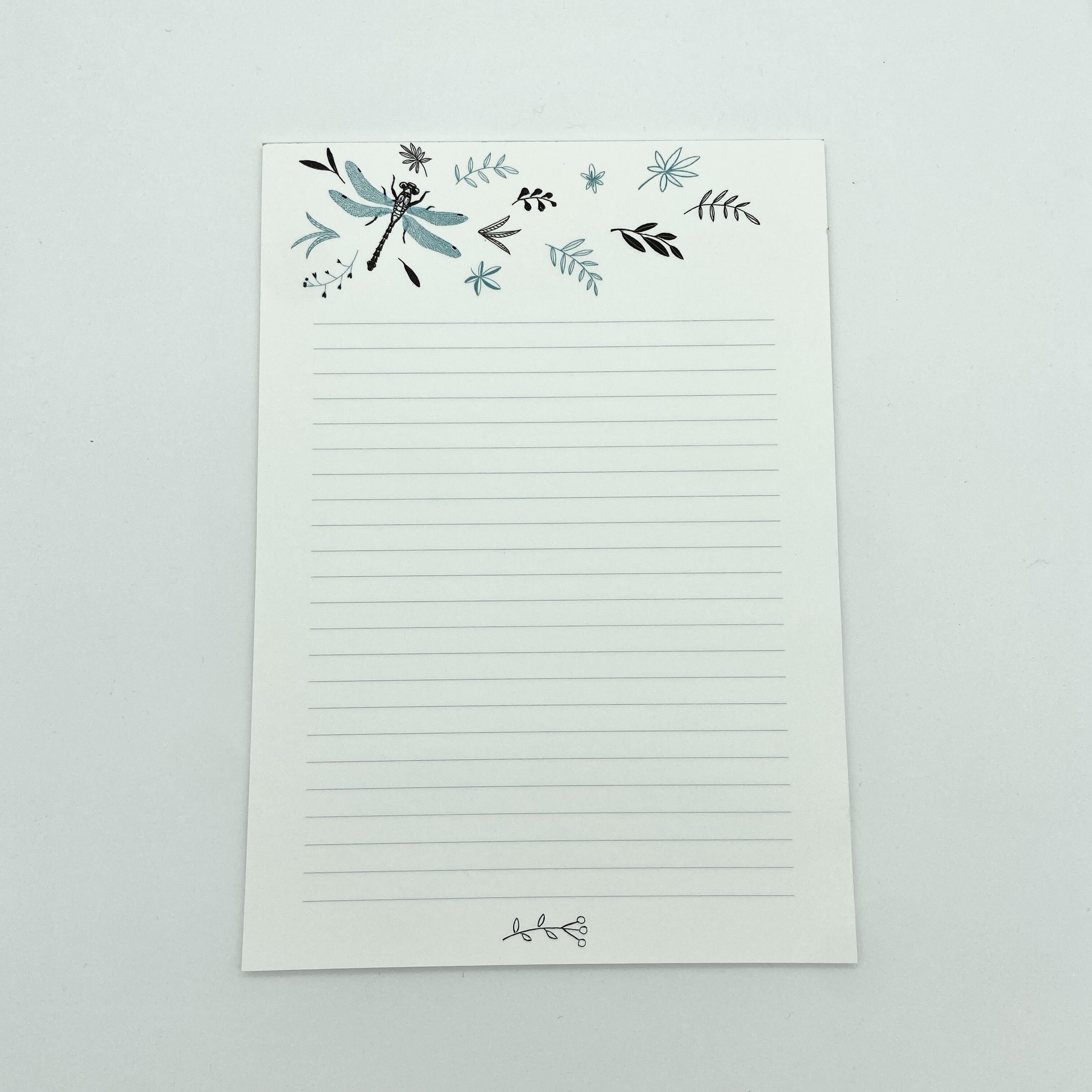 Ein gefütterter Notizblock mit einer blauen Libelle und einem Blumenmuster am oberen Rand und einem kleinen dekorativen Zweig am unteren Rand auf einem schlichten weißen Hintergrund.