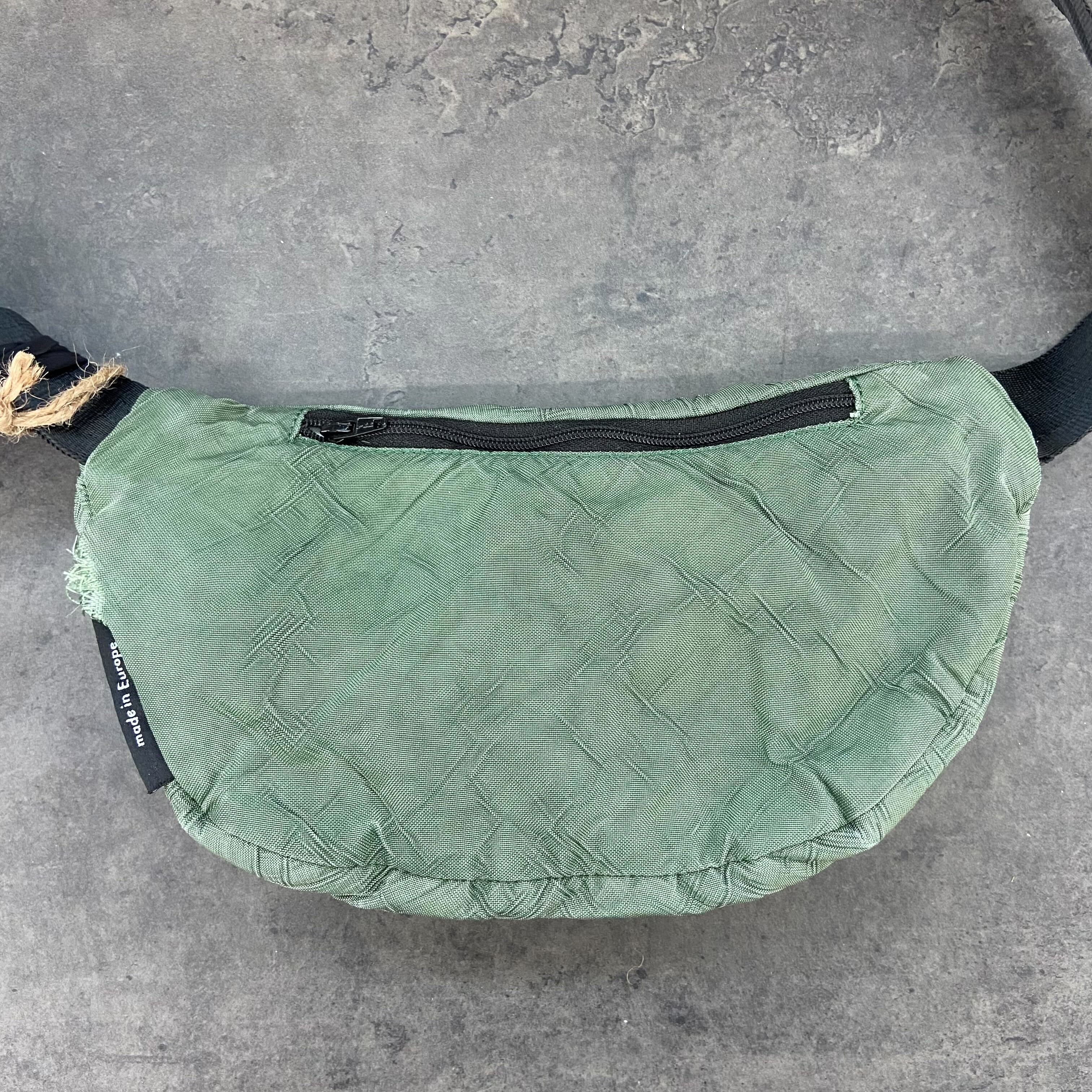 Grüne Bauchtasche mit Reißverschluss und Schnurdetail auf grauem Hintergrund.