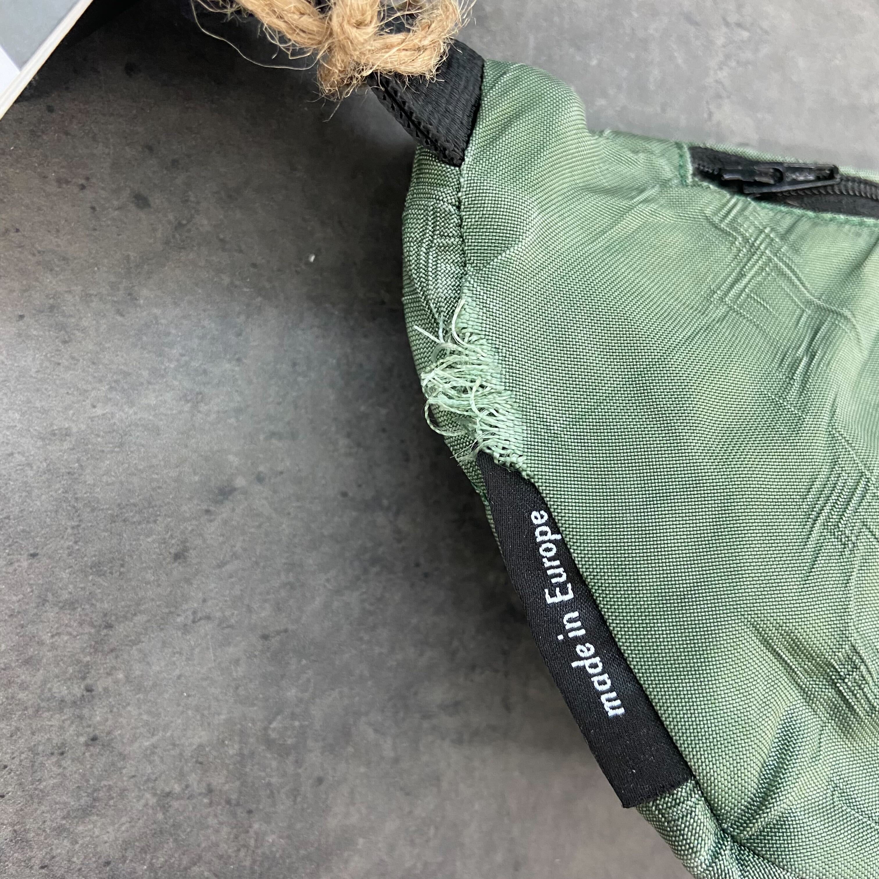 Grüner Rucksack mit ausgefranstem Rand, Etikett zeigt "made in Europe", auf grauem Hintergrund.