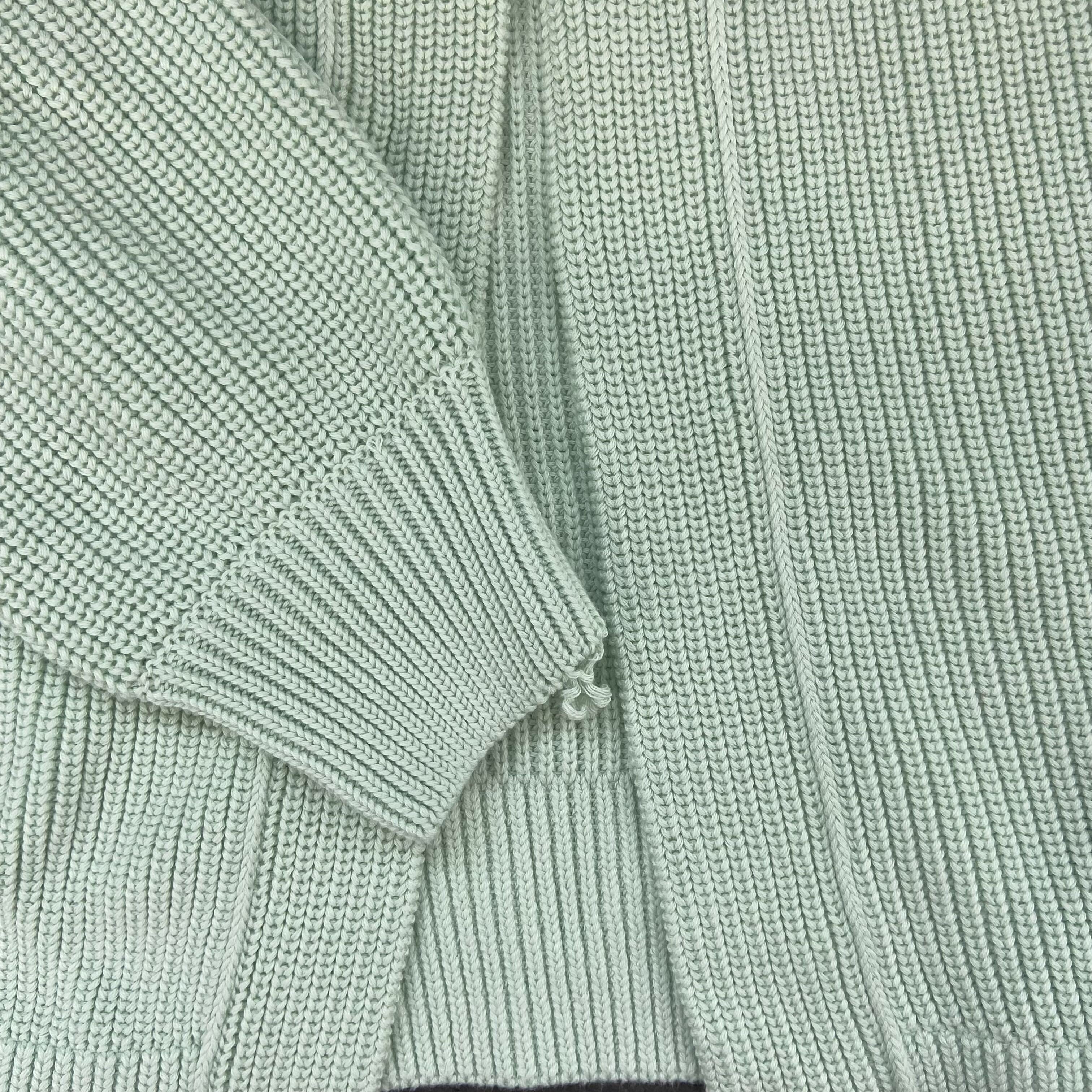 Grüngrauer Strickpullover aus grobem Garn, mit langen Ärmeln und Rippbündchen, auf einer flachen Oberfläche drapiert.