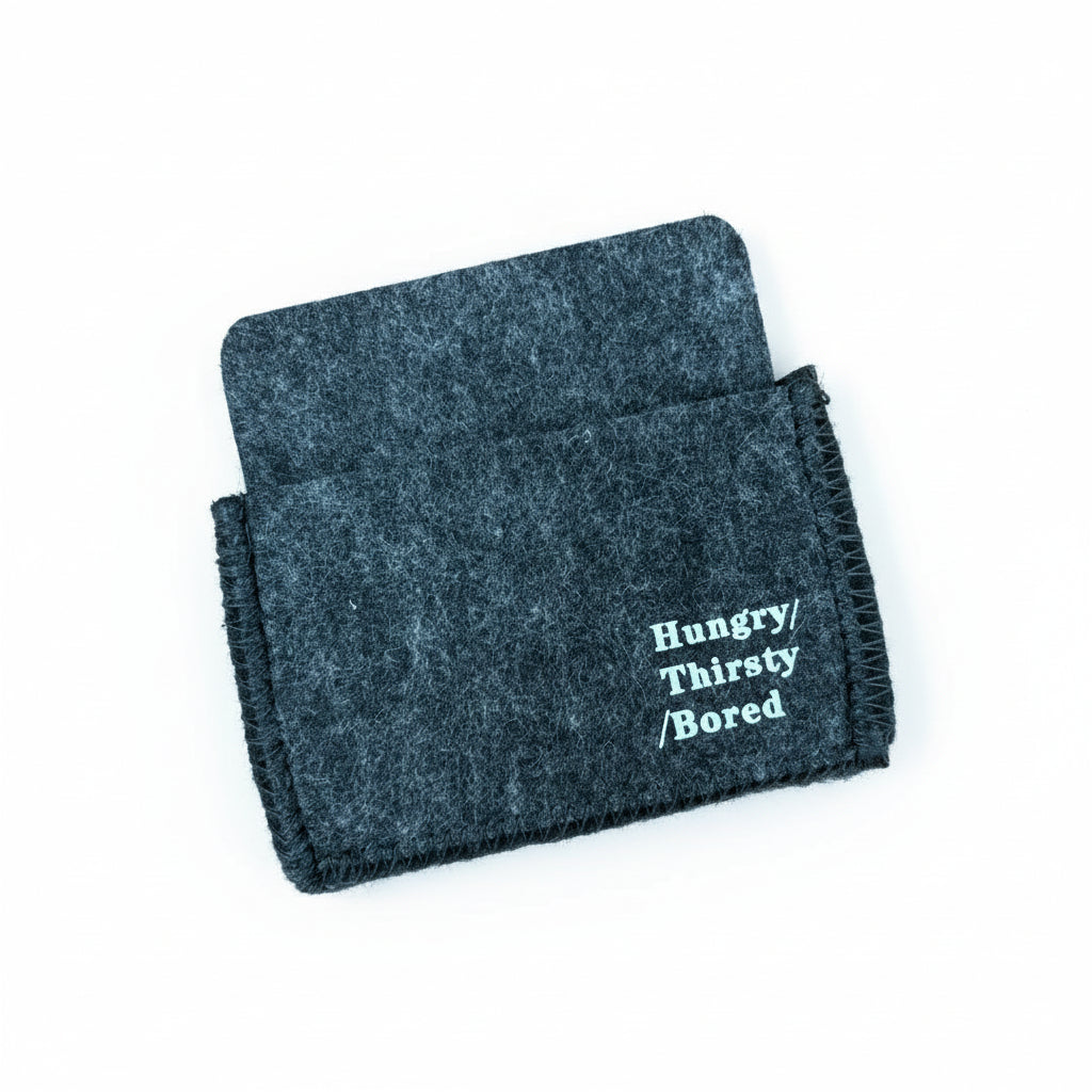 Ein kleiner, dunkelgrauer Filzbeutel mit schwarzen Nähten liegt auf einer Holzfläche. Auf der Vorderseite des Beutels steht in weißer Schrift "Hungrig/ Durstig/ Gelangweilt".