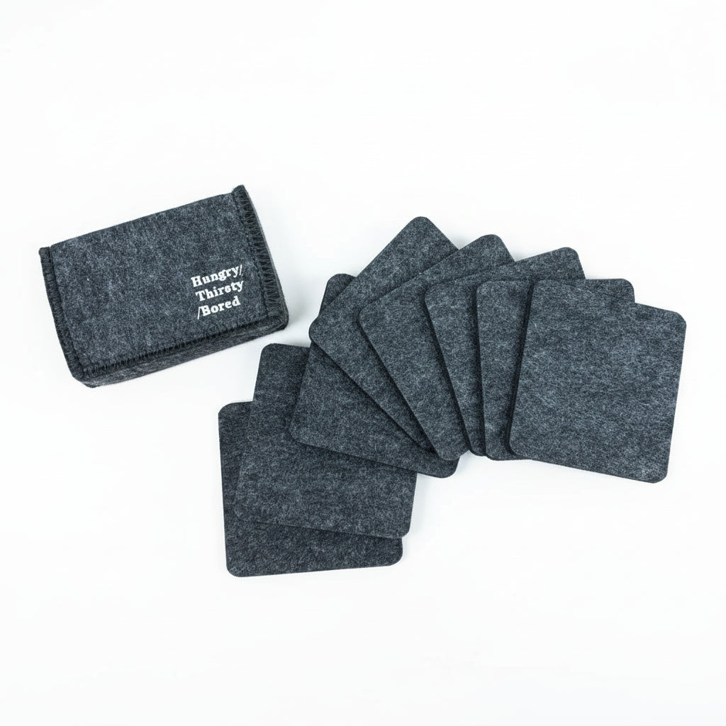 Ein Satz dunkelgrauer Filzuntersetzer liegt auf einem Holztisch neben einer passenden Filztasche mit der Aufschrift "Hungry Thirsty Bored" in weißer Schrift.