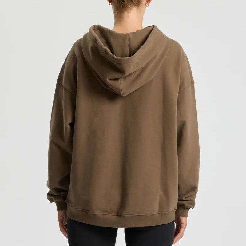 Eine Person mit blondem Haar in einem ordentlichen Dutt, die einen lockeren braunen Kapuzenpulli und schwarze Leggings trägt, steht mit dem Gesicht von der Kamera weg vor einem schlichten hellen Hintergrund.