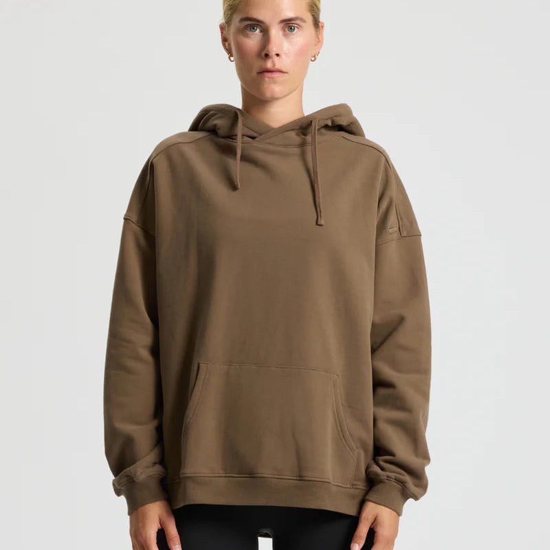 Eine Person mit zurückgebundenen blonden Haaren trägt einen lockeren braunen Kapuzenpulli und eine schwarze Hose, steht vor einem schlichten hellgrauen Hintergrund und schaut mit neutralem Blick direkt in die Kamera.