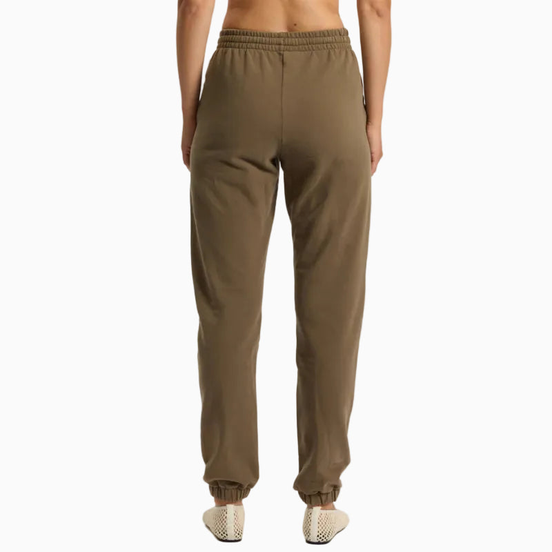 Eine Person steht mit dem Gesicht zur Wand und trägt einen braunen Sport-BH mit Riemen, eine passende braune Jogginghose und helle Slipper vor einem schlichten weißen Hintergrund.