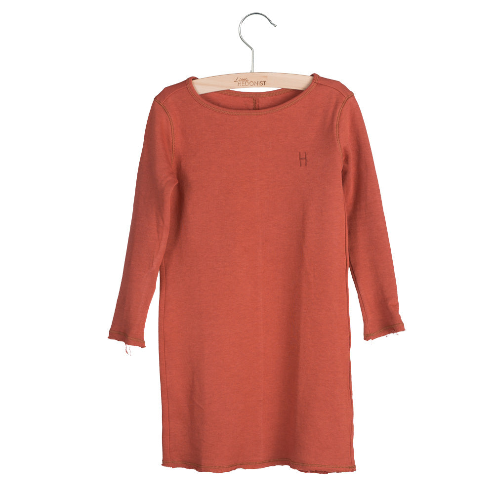 Ein langärmeliges, rostfarbenes Kleid für Kinder hängt an einem Holzbügel. Das Kleid hat ein einfaches Design mit rohen Kanten an den Ärmeln und am Saum und einem kleinen gestickten H auf der linken Brust.