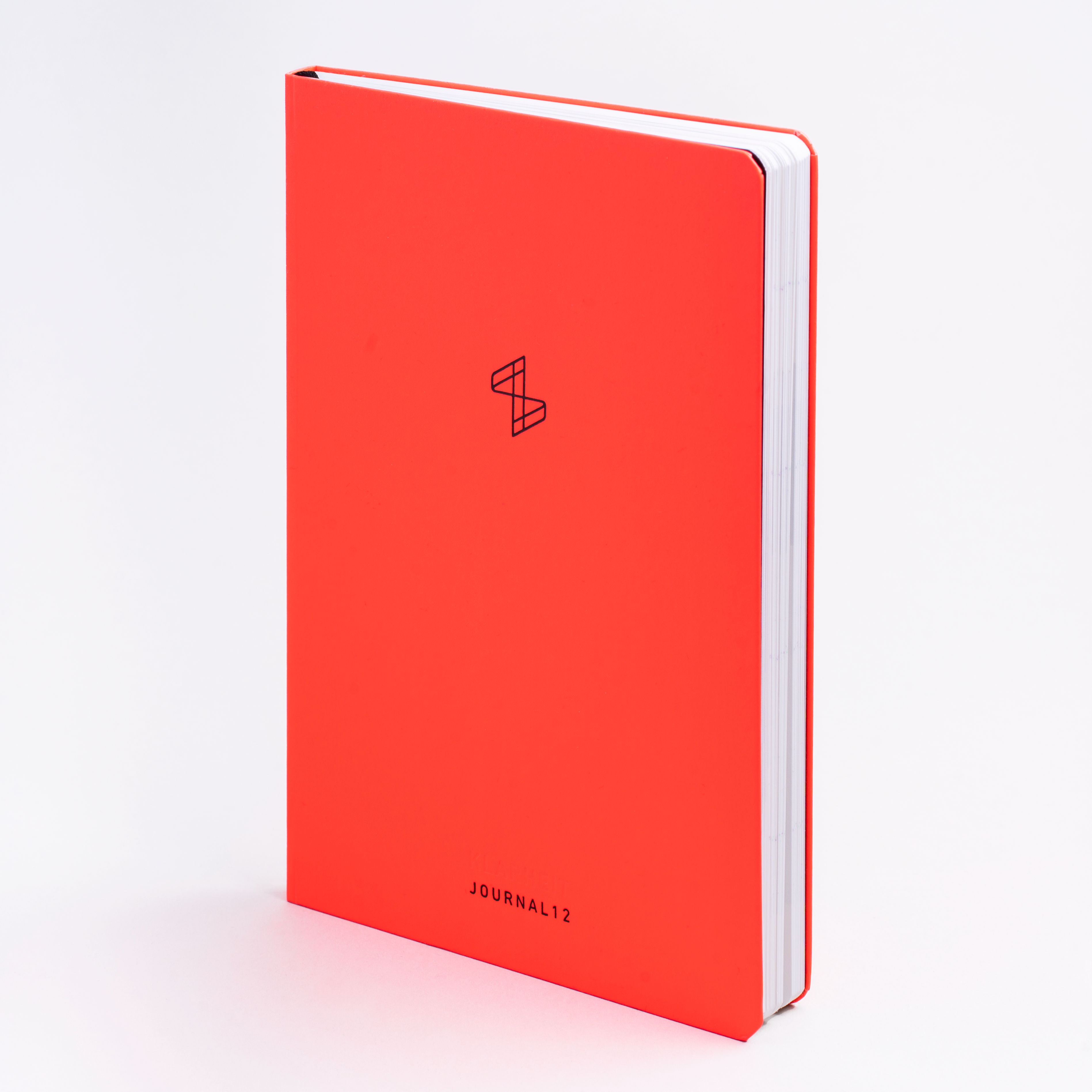 Rotes Notizbuch mit schlichtem geometrischen Logo und Text "JOURNAL12" auf einem hellen Hintergrund.