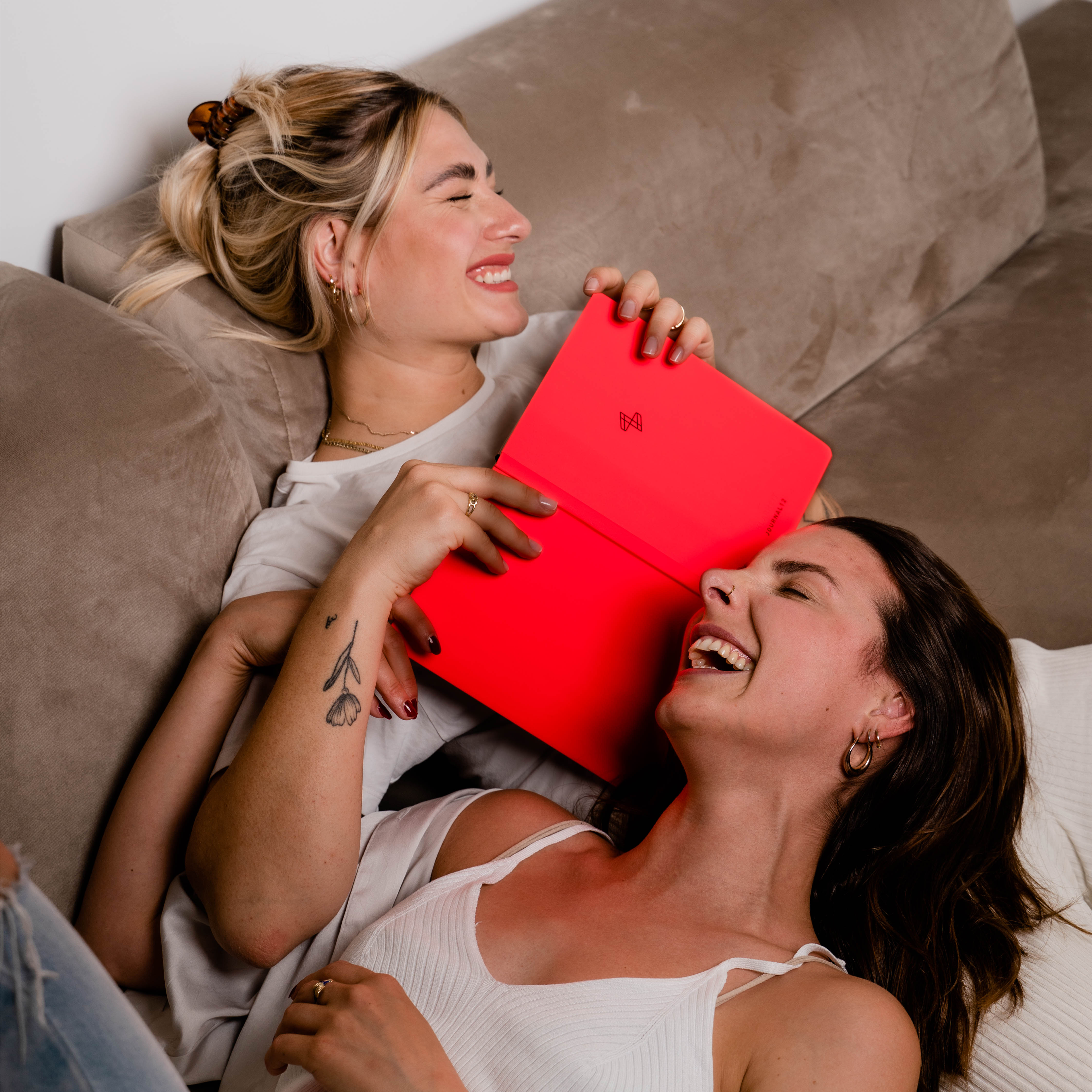 Zwei lachende Frauen liegen auf einem Sofa, eine hält ein rotes Notizbuch.