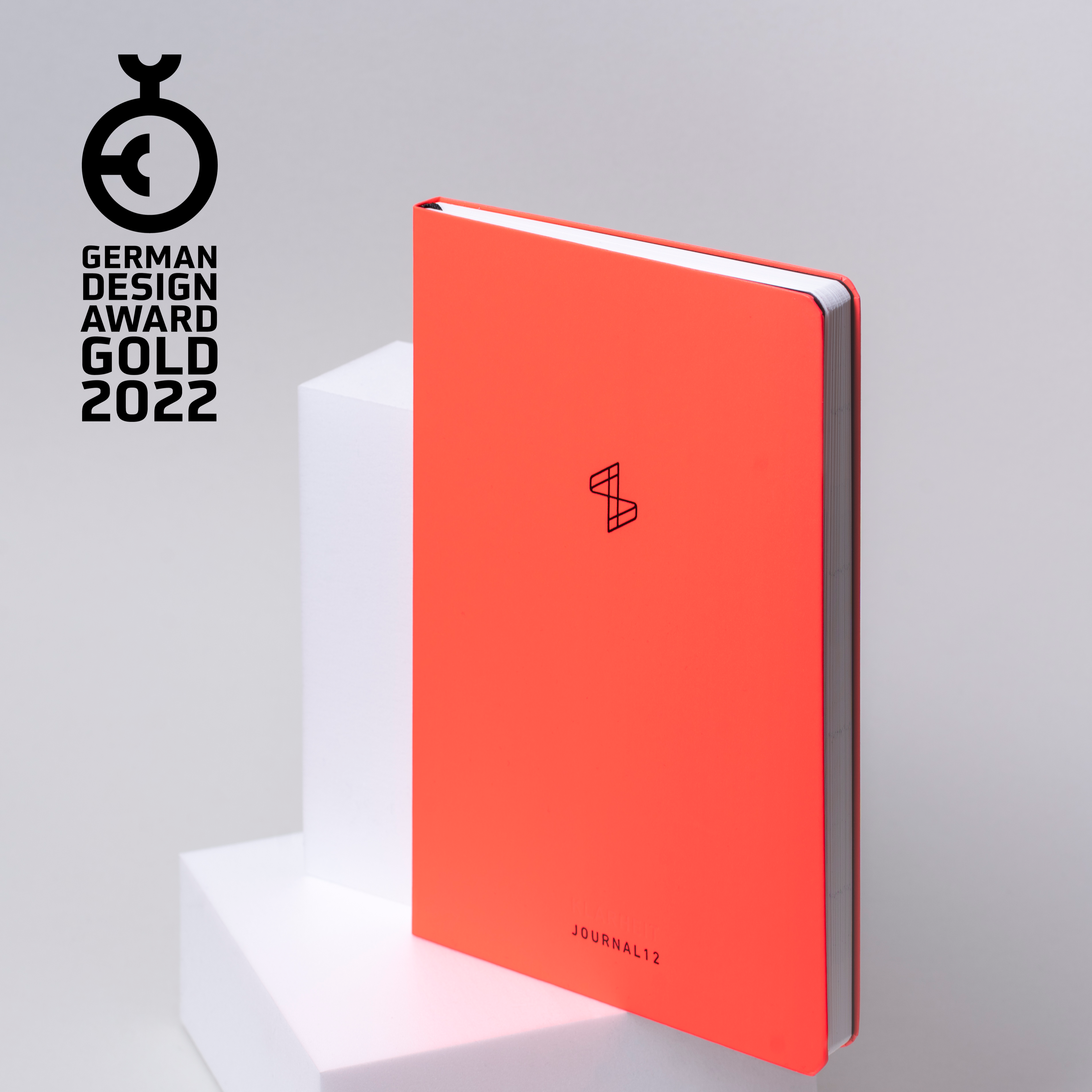 Rotes Notizbuch mit dem Logo des German Design Award Gold 2022, aufgestützt auf weiße Blöcke.
