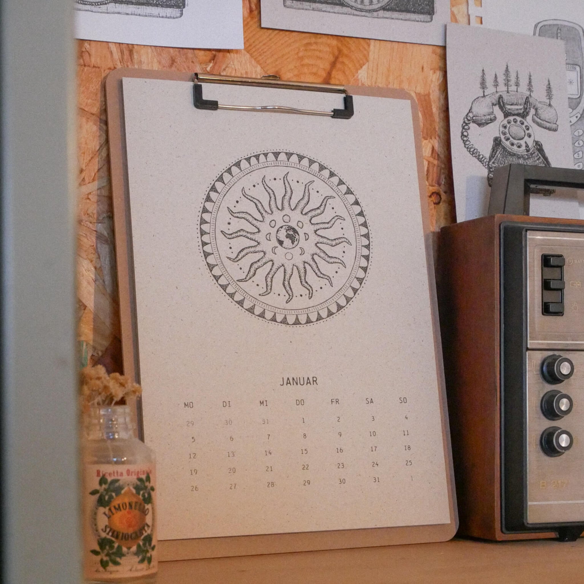 Ein Klemmbrett mit einem Januar-Kalender mit kreisförmigem Sonnenmotiv steht auf einem Holzregal neben einem alten Radio und einer kleinen Glasflasche mit einem Blumenetikett. Dahinter ist ein schwarz-weißes Kunstwerk an die Wand gepinnt.