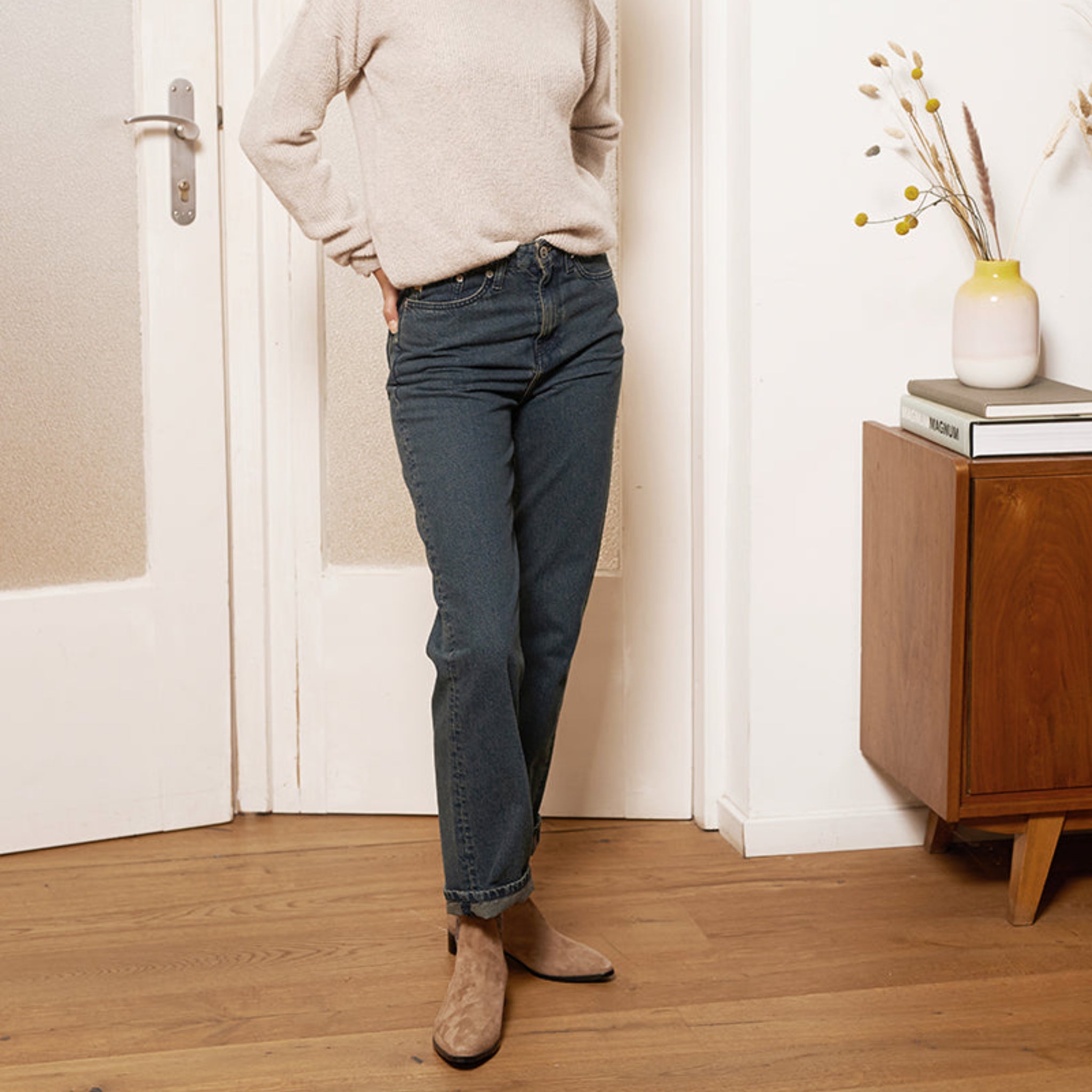 Person in beige Pullover und blauer Jeans steht neben einem Holzmöbel mit Vase und Büchern auf Holzfußboden.