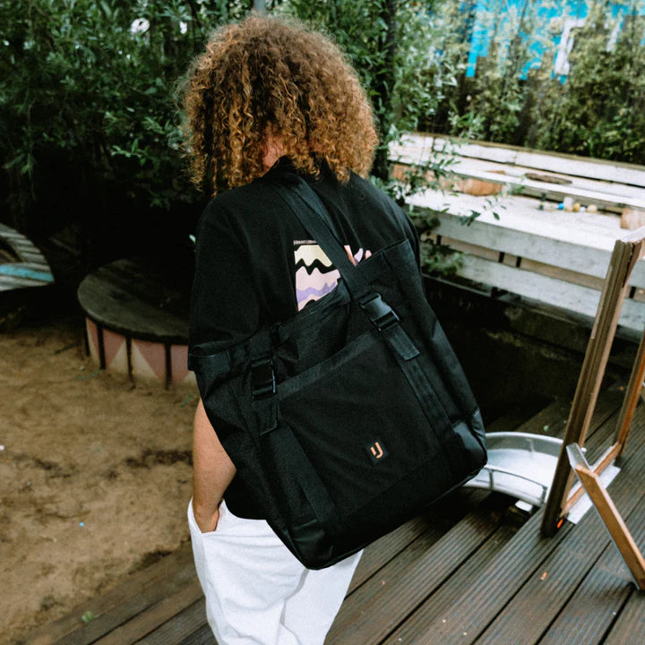 Eine Person mit lockigem Haar trägt ein schwarzes T-Shirt mit einem bunten Muster und hat eine große schwarze Tasche über der Schulter.