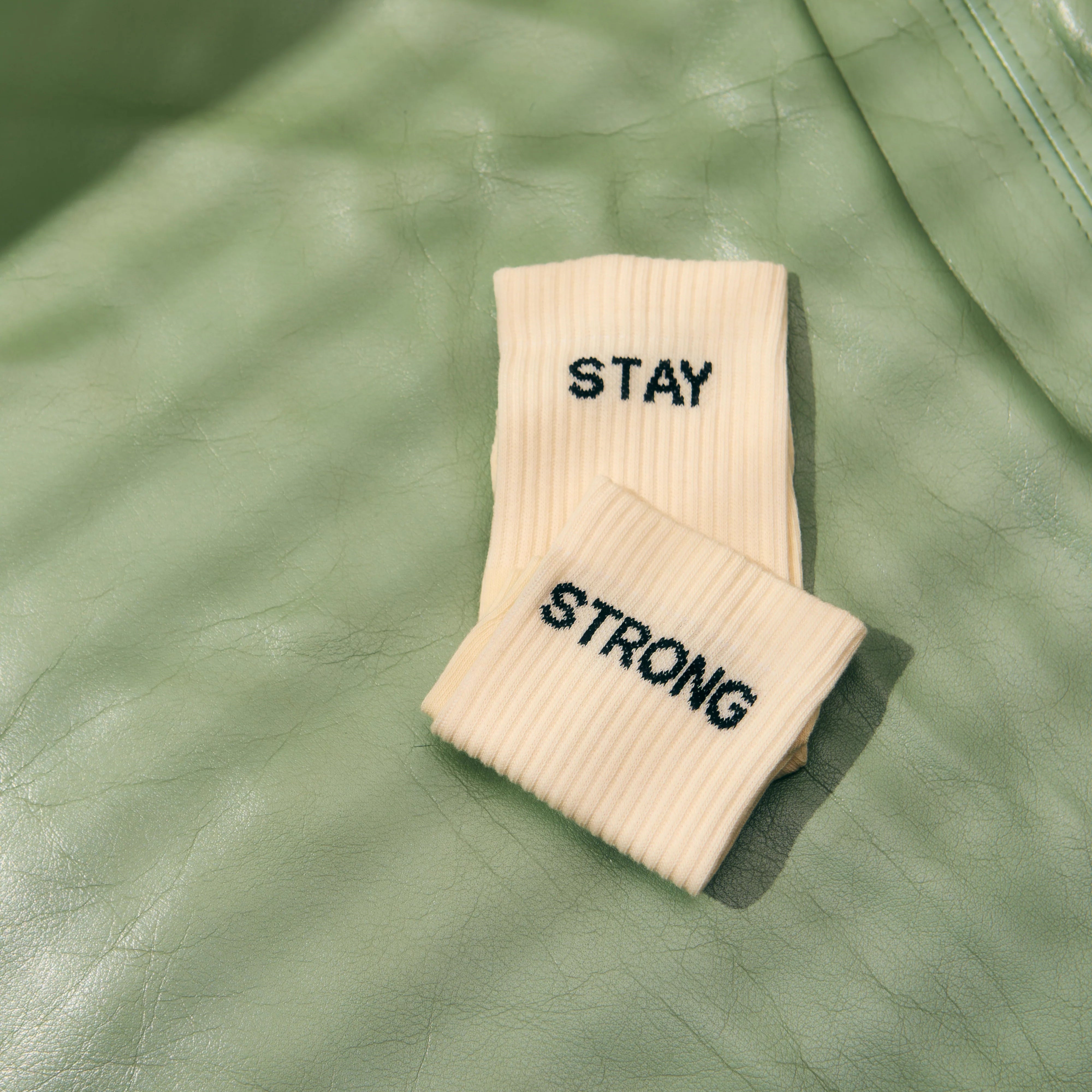 Zwei cremefarbene Rippensocken mit den Aufschriften STAY und STRONG in schwarzer Schrift liegen auf einer weichen, hellgrünen Lederoberfläche.