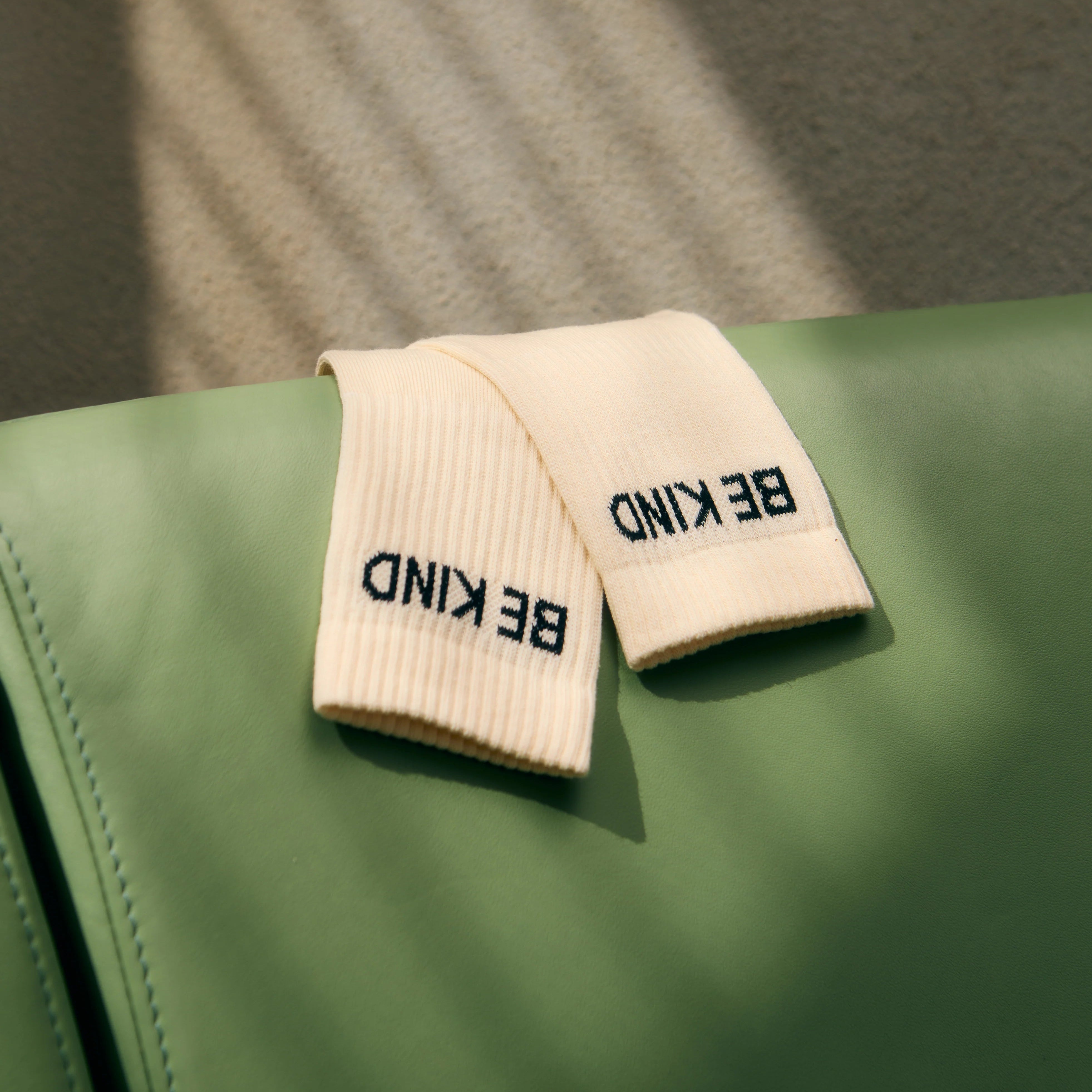 Ein Paar beigefarbene Socken mit der Aufschrift BE KIND (Sei freundlich) sind auf einer grünen Fläche platziert, wobei die Schatten der Jalousien auf den Hintergrund geworfen werden.
