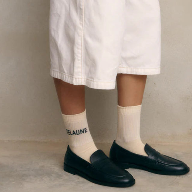 Eine Person in weißen, knielangen Shorts, cremefarbenen Socken mit der Aufschrift TELAUNE und schwarzen Halbschuhen steht auf einem Betonboden vor einer schlichten Wand.