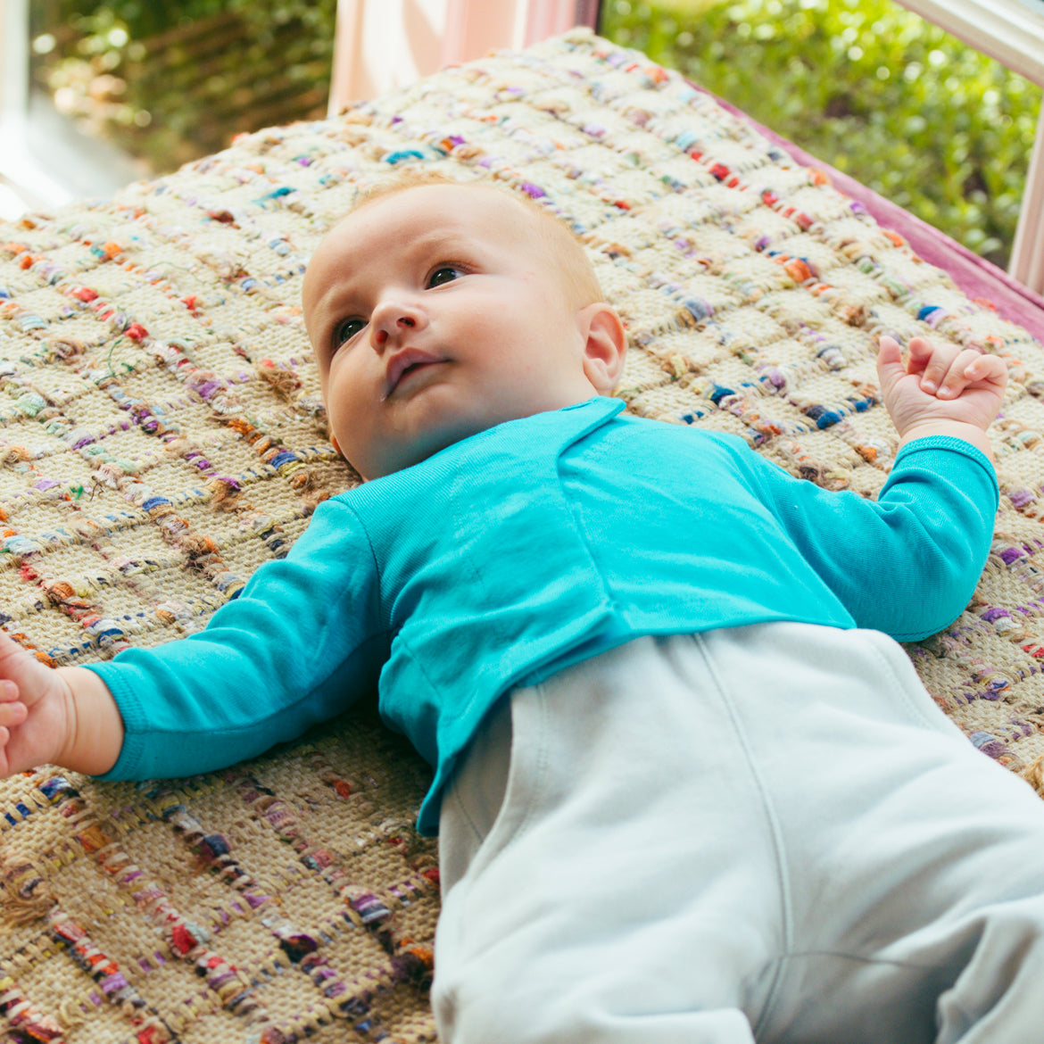 Ein Baby in einem blauen Oberteil liegt auf einer bunten Decke und schaut zur Seite.