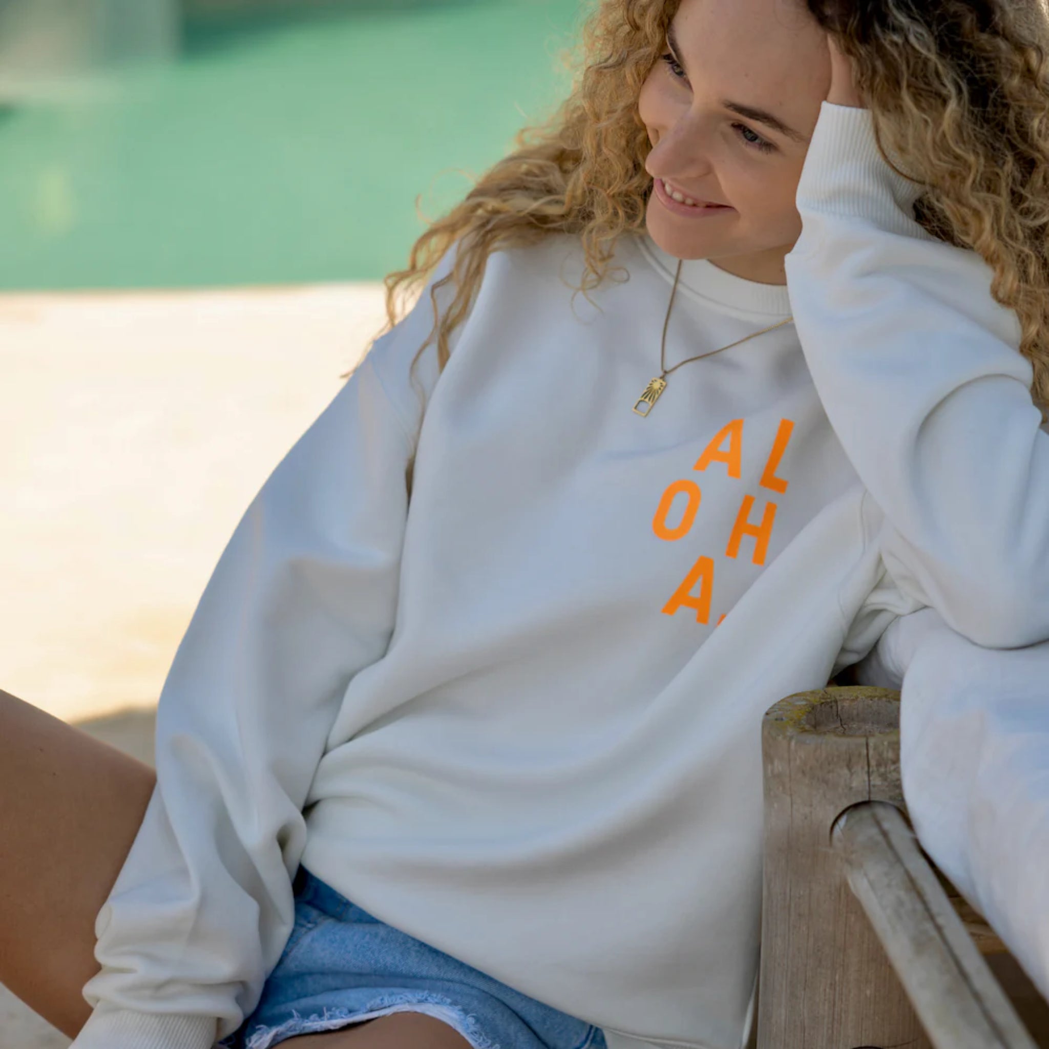Eine lächelnde Frau mit lockigem Haar trägt ein weißes Sweatshirt mit orangefarbenem ALOHA-Aufdruck und Jeansshorts. Sie sitzt im Freien am Wasser und stützt ihren Kopf auf ihre Hand.