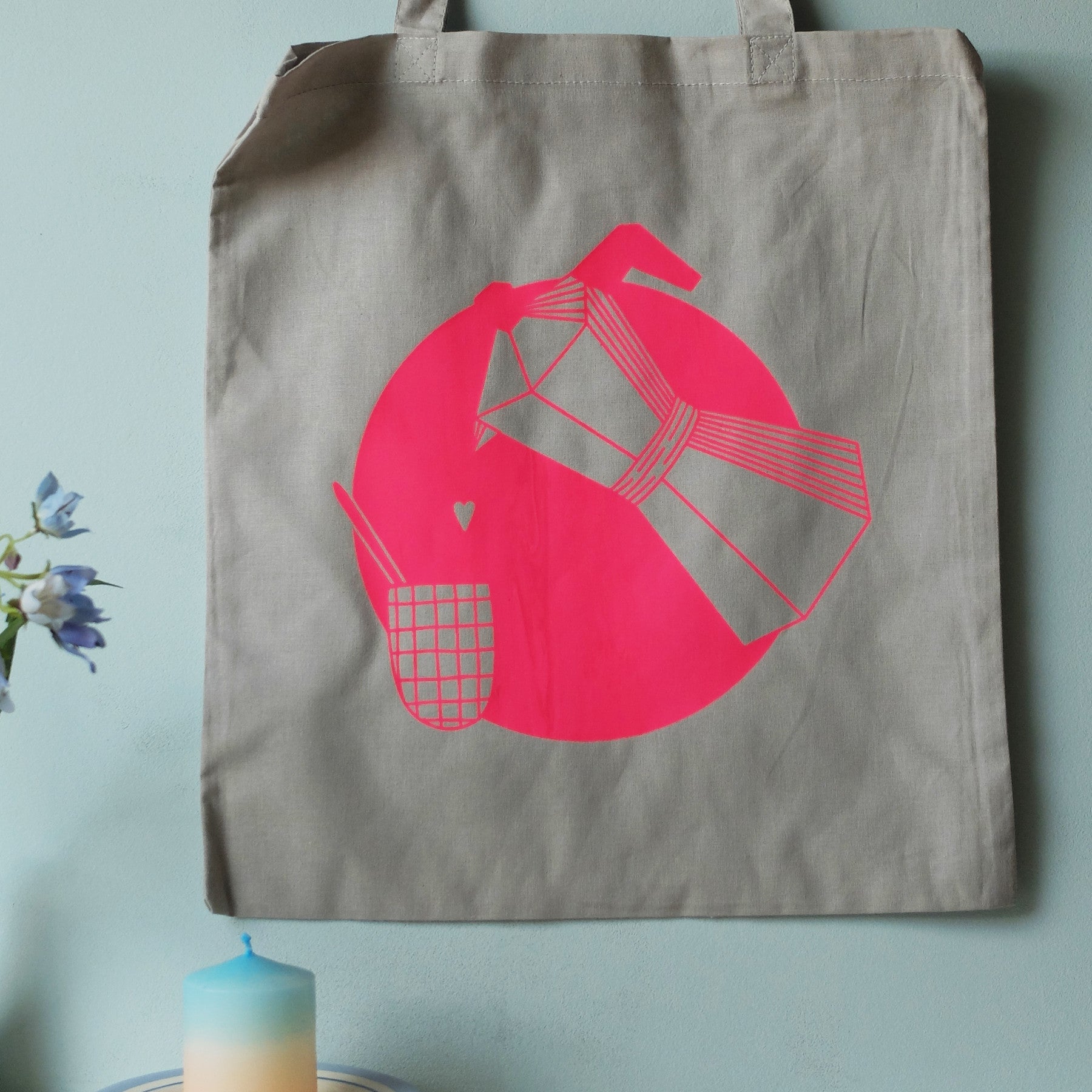 Eine graue Tragetasche mit rosafarbener Grafik, die eine gefaltete Serviette, ein kleines kariertes Täschchen und ein Herz zeigt, alles vor einem rosafarbenen kreisförmigen Hintergrund. Die Tasche hängt an einer blauen Wand neben Blumen und einer blauen Kerze.