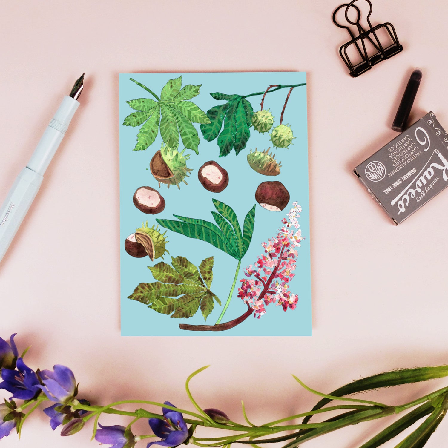 Eine Postkarte mit botanischen Illustrationen von Blättern, Blumen und Samen auf blauem Hintergrund. Sie liegt auf einer rosa Fläche mit einem Stift, einer Büroklammer, einem Radiergummi und violetten Blumen in der Nähe.