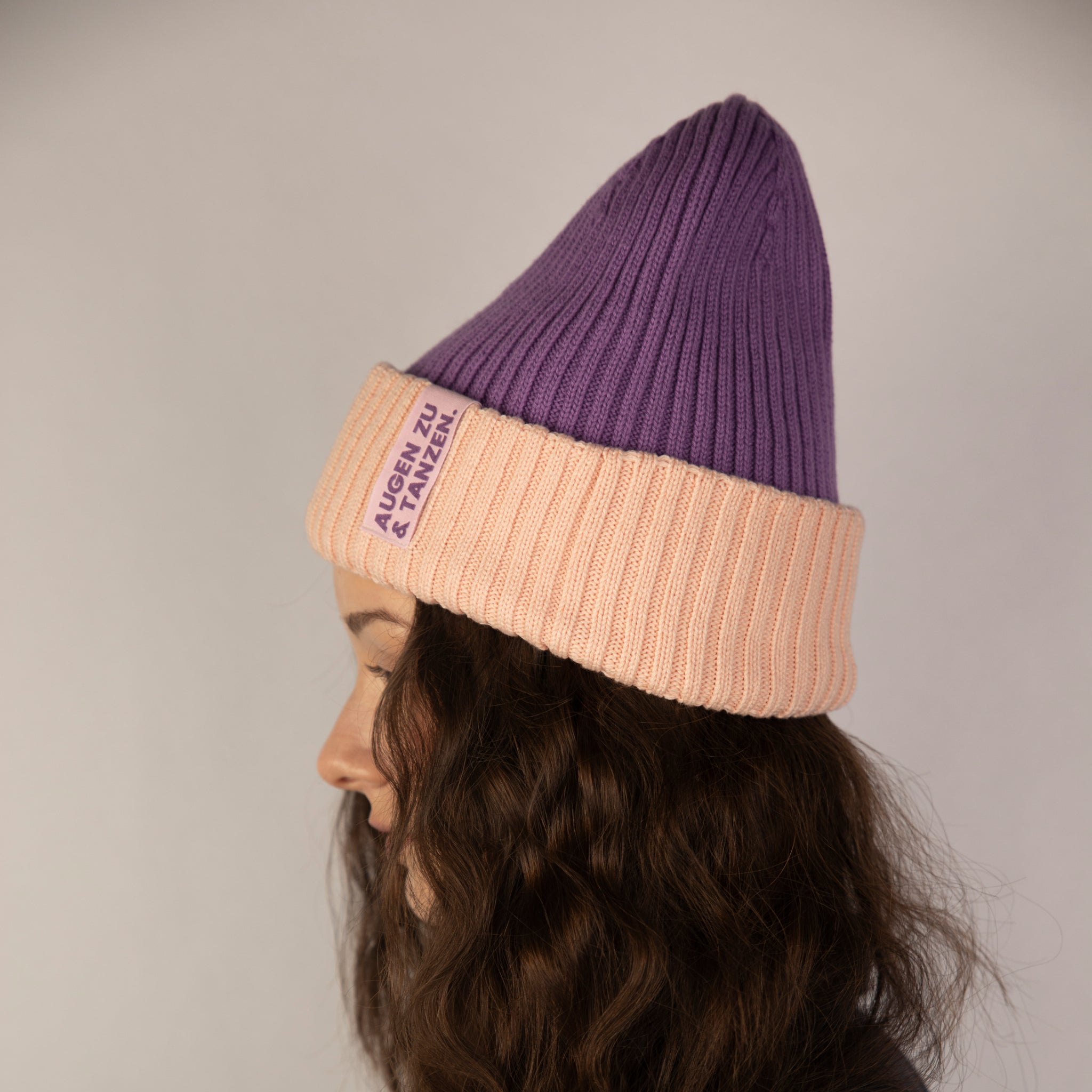 Eine Person mit gewelltem braunem Haar ist im Profil abgebildet und trägt eine gerippte violette Mütze mit breitem, umgeschlagenem pfirsichfarbenem Rand und einem violetten Etikett darauf. Der Hintergrund ist schlicht und neutral.