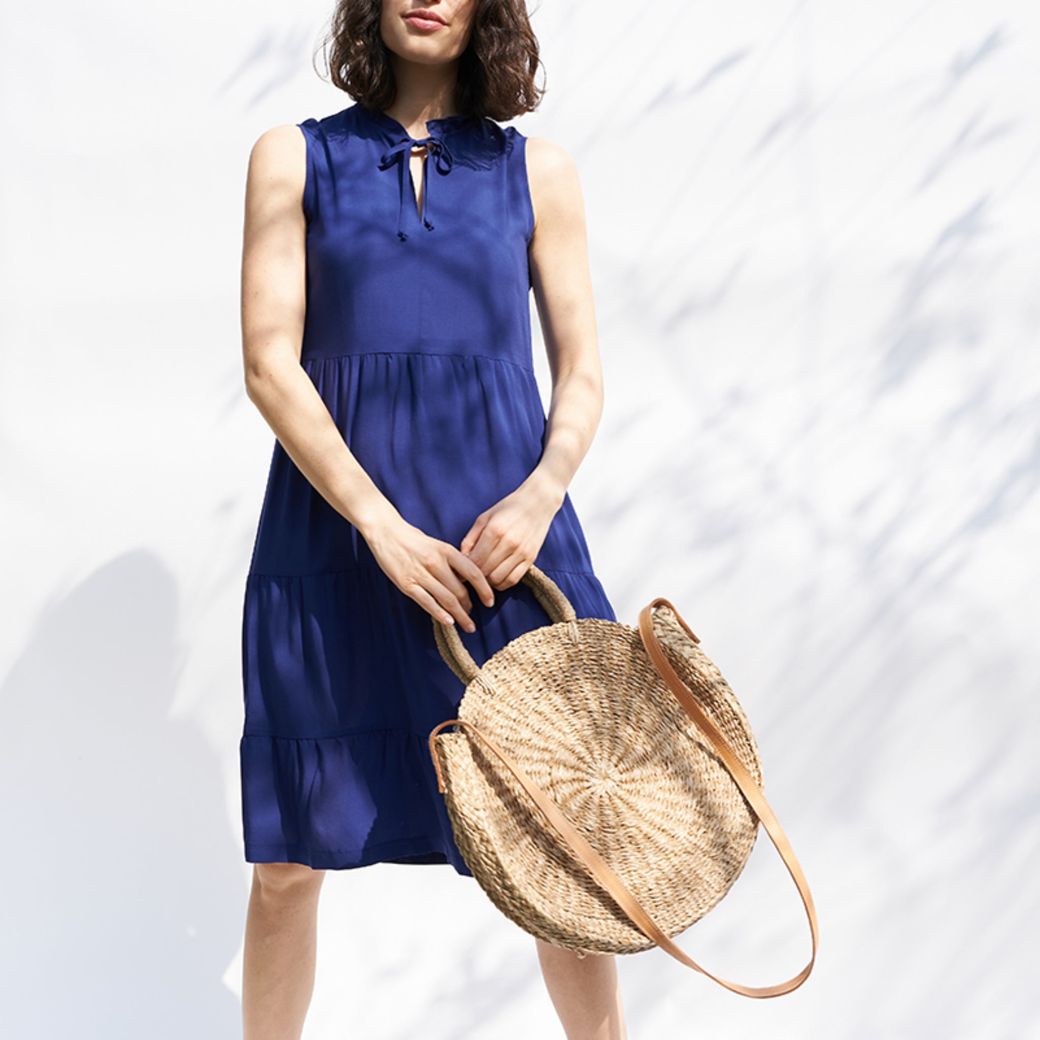 Frau in einem blauen ärmellosen Kleid hält eine runde Strohtasche vor einem weißen Hintergrund.