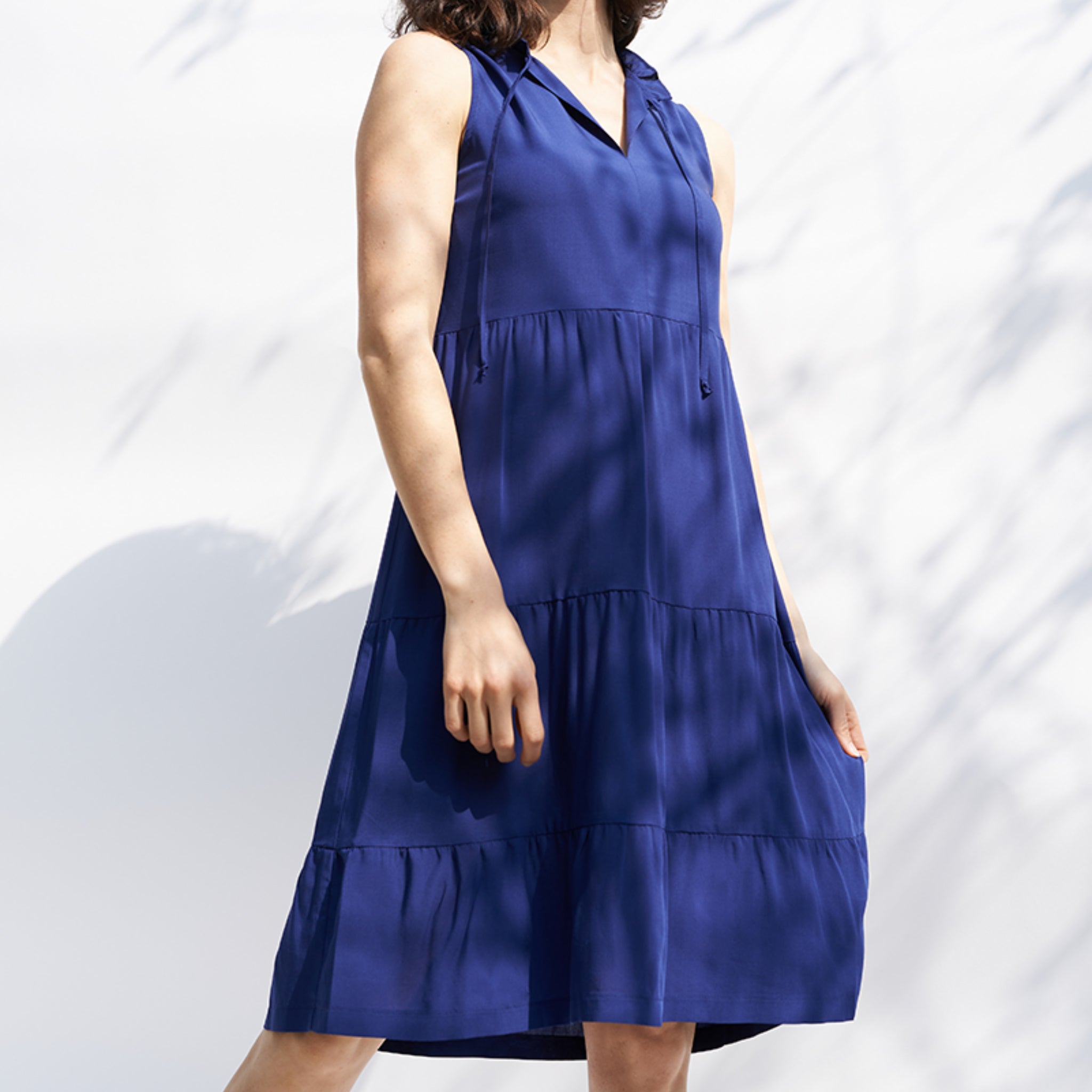 Frau in einem blauen, ärmellosen Kleid steht vor einer weißen Wand mit Schatten.