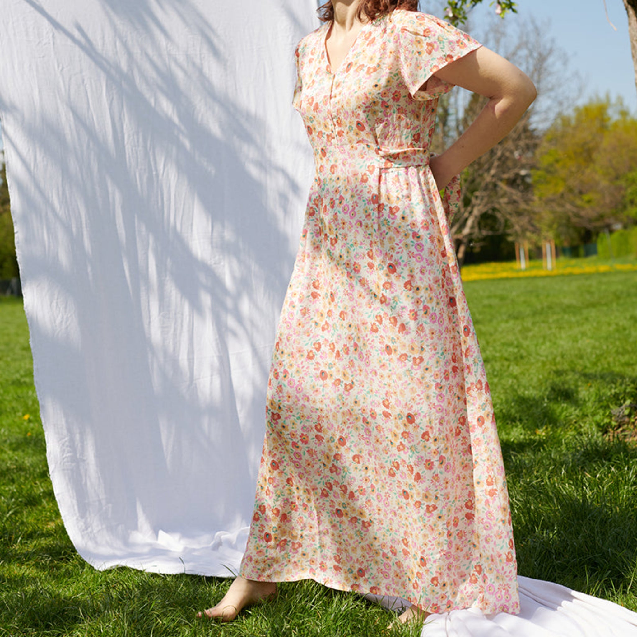 Eine Person trägt ein langes, geblümtes Kleid und steht barfuß auf einer Wiese neben einem weißen Hintergrund.