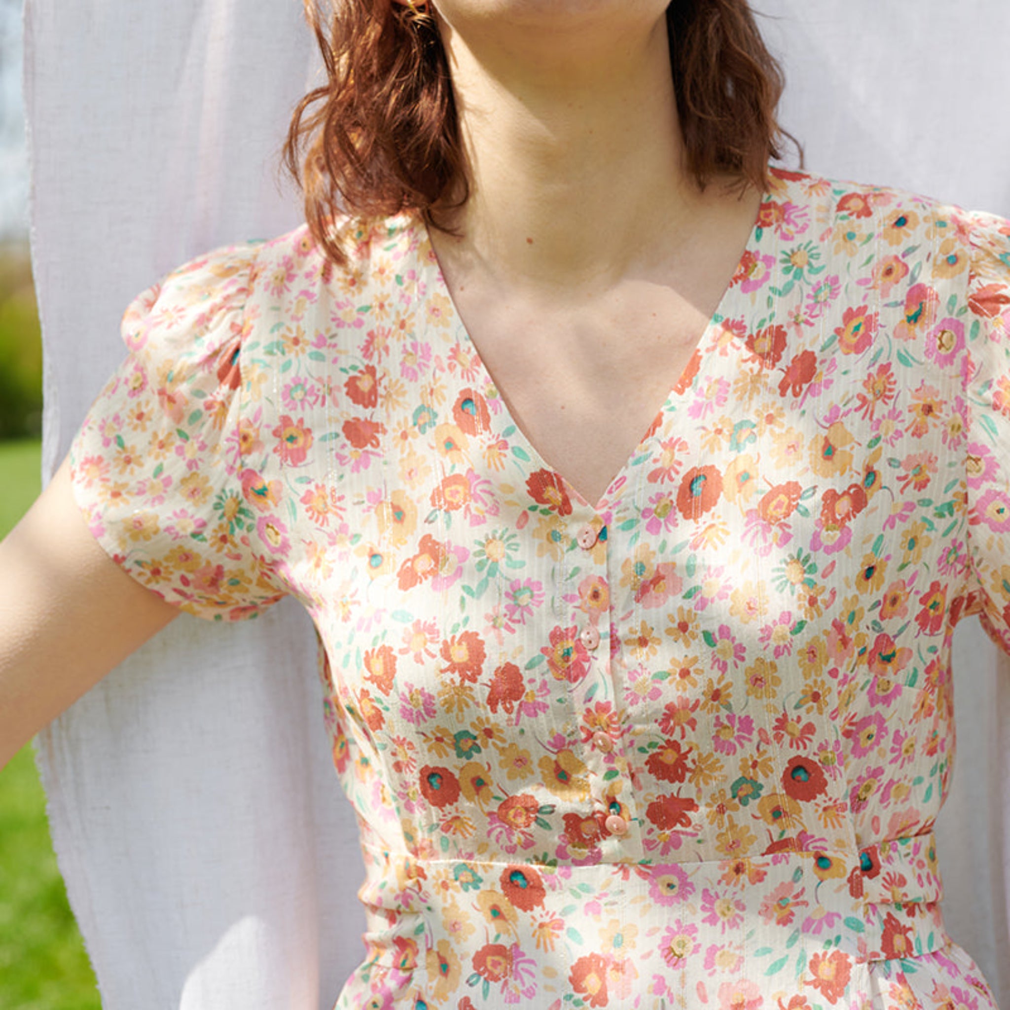 Eine Frau trägt ein blumiges Kleid mit V-Ausschnitt im Freien. Ihr Gesicht ist nicht sichtbar.