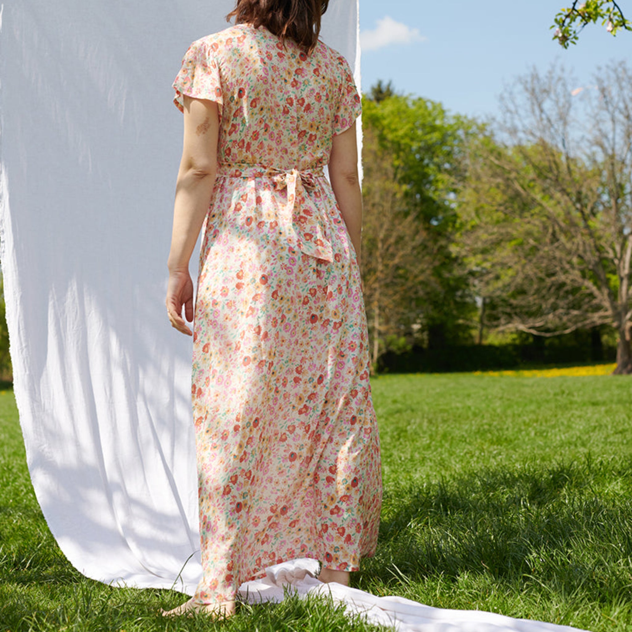Eine Person in einem geblümten Kleid steht im Freien auf einer Wiese, vor einem weißen Hintergrundtuch.