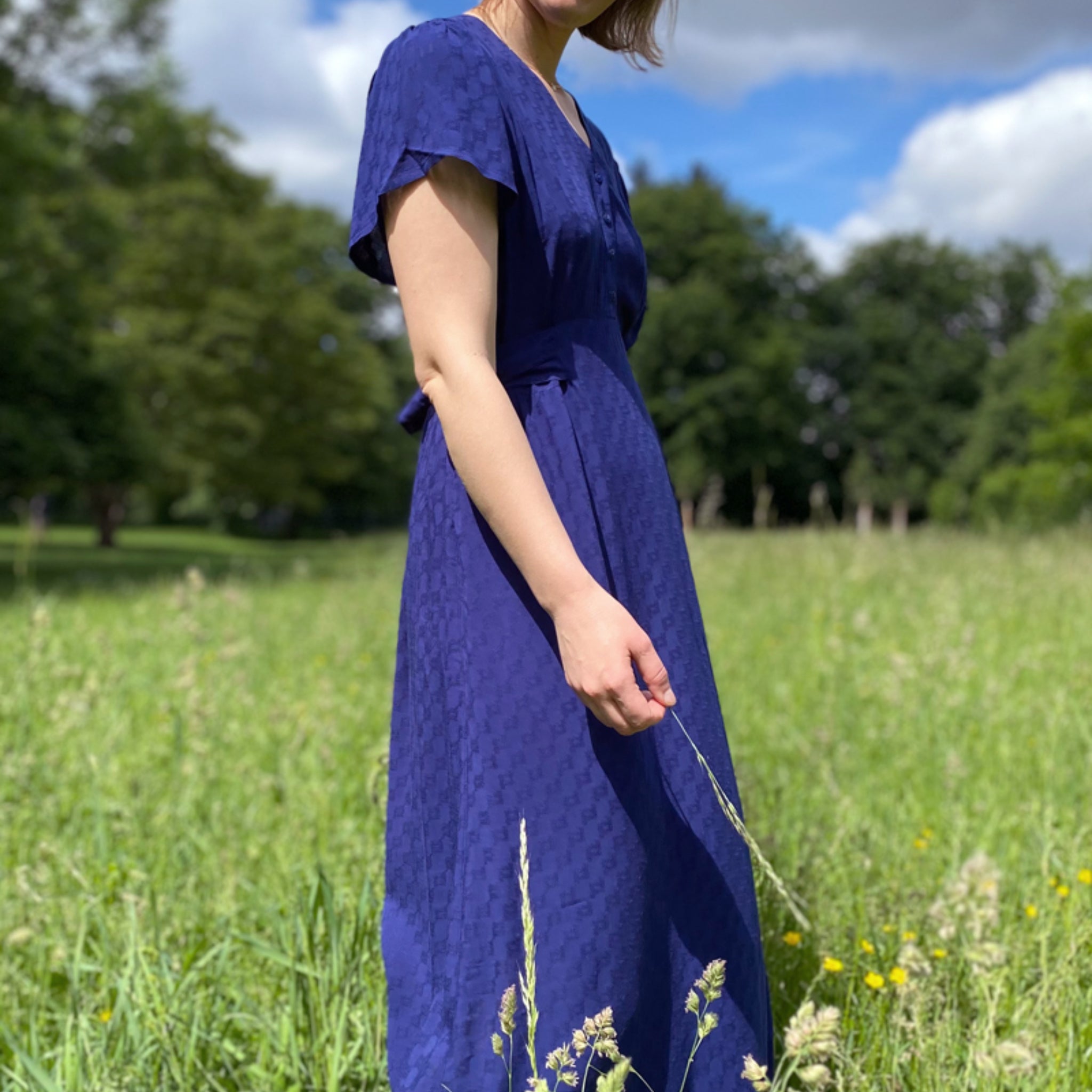 Person in einem blauen Kleid steht auf einer Wiese mit Bäumen im Hintergrund, bei sonnigem Wetter.