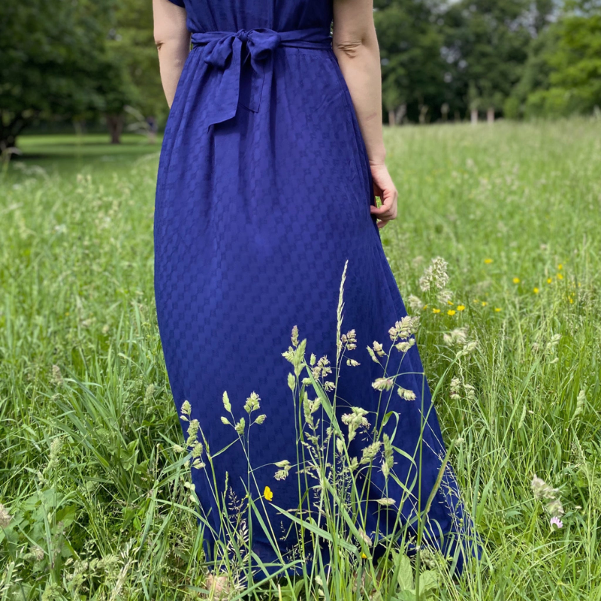 Person von hinten in einem blauen Kleid steht auf einer Wiese mit hohem Gras.
