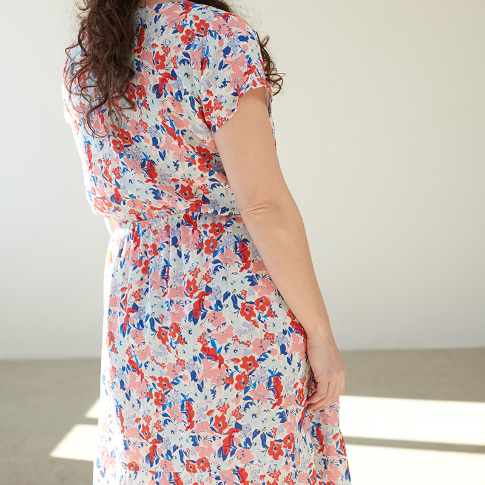 Frau in einem weißen Kleid mit buntem Blumenmuster, Rückenansicht, steht in einem hellen Raum.